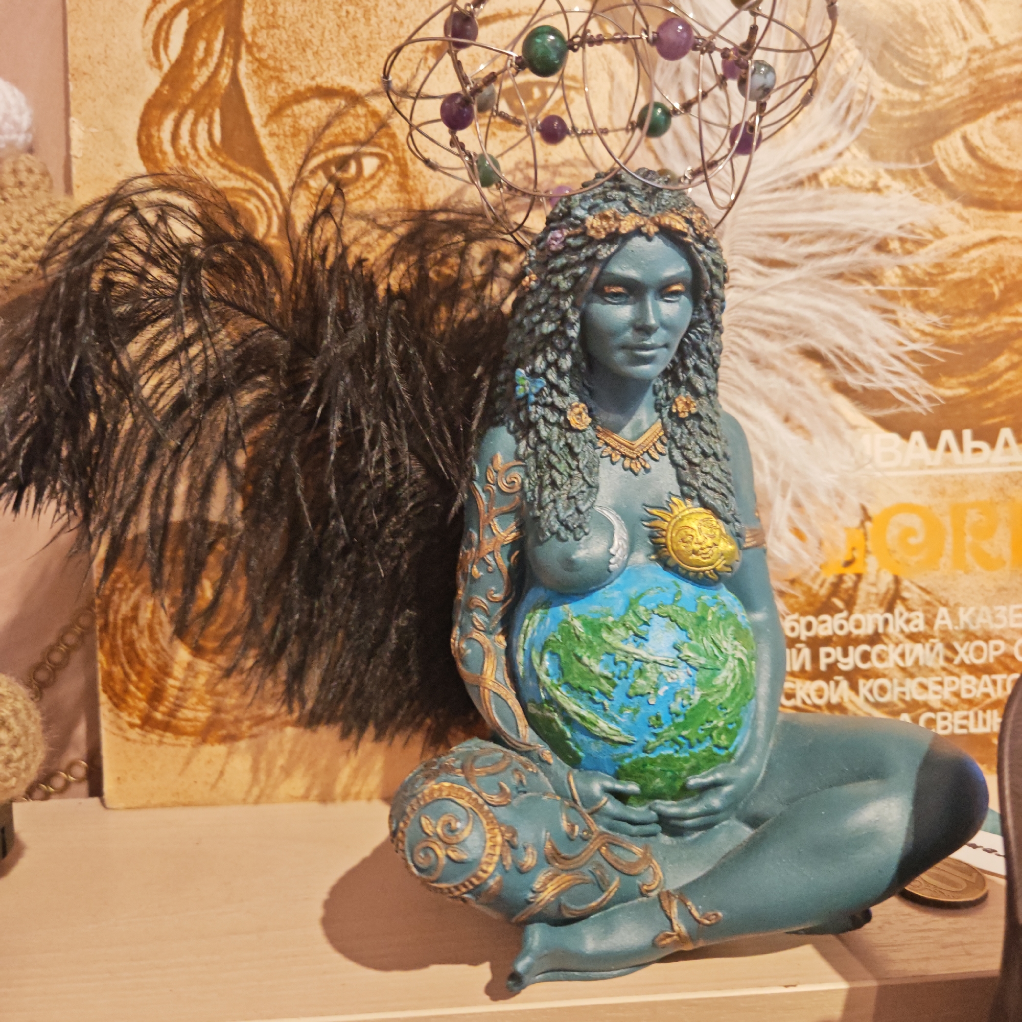 A photograph shows a teal-colored statuette of a pregnant woman sitting cross-legged and holding a globe on her abdomen. The statuette has gold-colored details, including a sun emblem on her chest, a necklace, and ornate designs on her arms and legs. Her hair is styled with leafy details and flowers, and she is topped with a spherical crown of wires and beads.
Behind the statuette is a collection of items, including black and white feather boas, a knitted object, and a printed poster with Russian text. The photograph appears to be taken indoors on a light-colored surface.
The printed poster with Russian text suggests a location in Russia, potentially Rostov, as asked by the request.