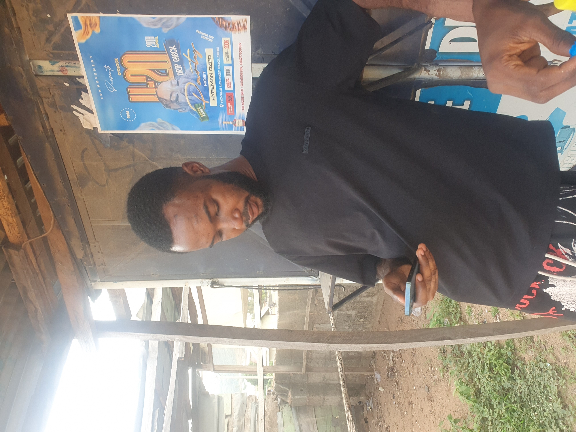 The image depicts a man holding a smartphone, standing beside a poster advertising an event called "H20 Drip Check" hosted by Hypeman Ozed at Royale Lounge Poolside on April 20th at 9 PM. The poster also lists ticket prices: 50K, 100K, and 200K. The man has a beard and wears a black t-shirt and patterned pants. The surroundings suggest an informal structure with wooden elements, and vegetation can be seen in the background. While the exact location within Lokoja, Nigeria, cannot be determined solely from the image, the event's locale is specified as Royale Lounge Poolside.
