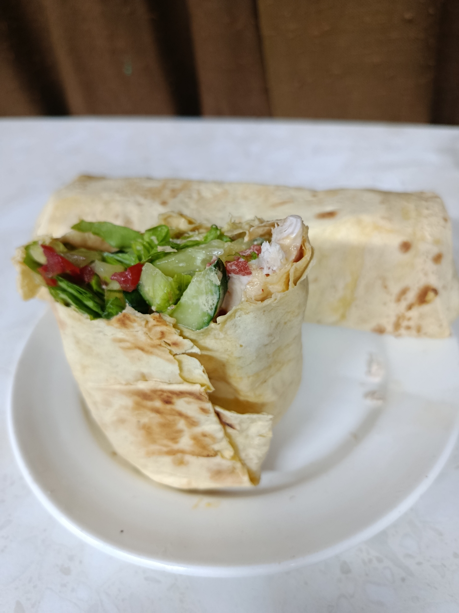 The image displays two food wraps, likely shawarma or burritos, presented on a white round plate. The foreground wrap stands upright and is partially unwrapped, revealing its fresh ingredients. Visible inside are pieces of green lettuce, sliced cucumbers, diced red vegetables (possibly tomatoes or bell peppers), and white pieces of protein, which appear to be chicken, all coated in a creamy, light-colored sauce. The flatbread used for the wraps is light beige with scattered browned, toasted marks.
Behind the prominent wrap, a second, fully rolled wrap lies horizontally on the plate, partially out of focus. The plate itself rests on a white surface that has a subtle, light grey speckled or marbled pattern. The background is completely blurred, showing a solid, warm brown color, suggesting an indoor setting like a kitchen or dining area. There are no people visible, and no text or brand names are discernible. The overall impression is of a freshly prepared meal, possibly homemade, presented simply and ready to be eaten. The time of day and specific location are not indicated by any cues in the image.