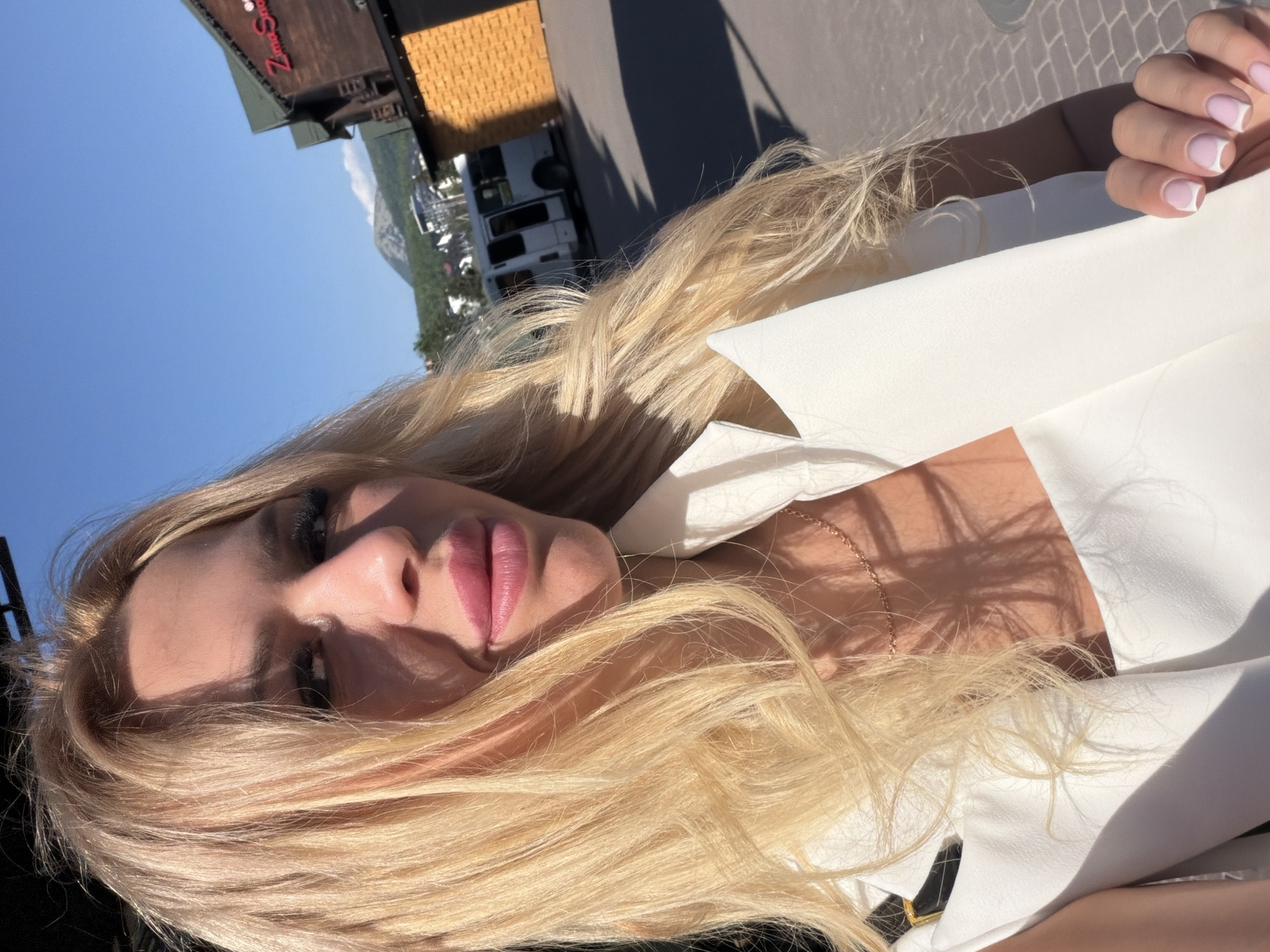 A close-up, selfie-style shot features a woman with blonde hair against a bright blue sky and a blurred background. She is wearing a white outfit, possibly a blazer, with a delicate gold chain visible around her neck. Her makeup includes mascara and pink lipstick. Her right hand, with neatly manicured nails, is positioned near her face.
In the background, elements of a setting suggest an outdoor location. A building with a sign that appears to read "Zima Snow" is visible, along with what looks like a white van or bus. The presence of trees and mountainous terrain further hints at a location like Polianytsia, Ukraine, possibly a ski resort area. The sunlight casts distinct shadows, indicating it is daytime, likely with clear weather. The overall mood conveyed is relaxed and casual.