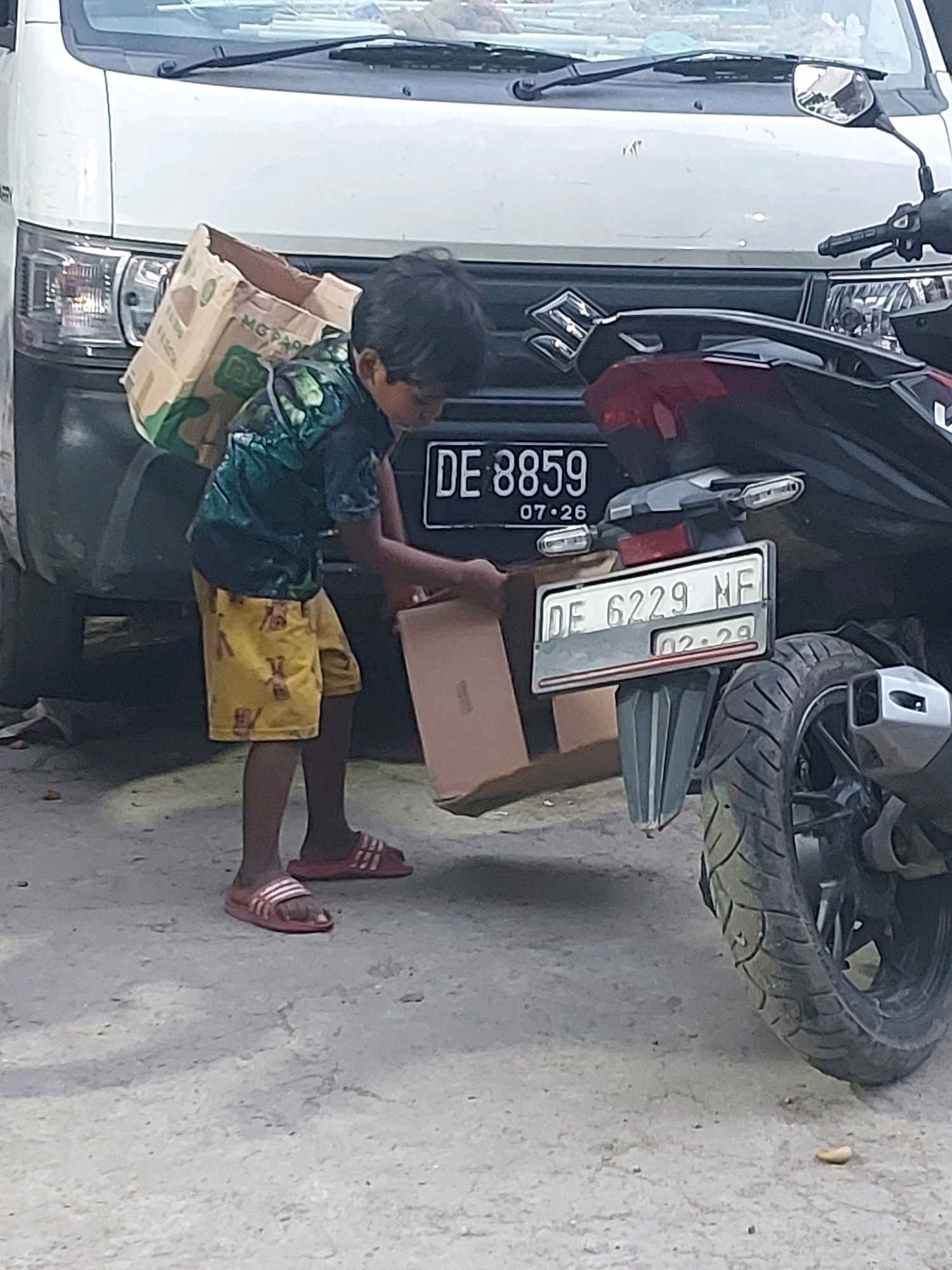 A young boy, likely of Indonesian ethnicity, is standing in front of a white van and a dark-colored motorcycle. He is wearing a dark green t-shirt with a camouflage pattern and yellow shorts with a repeating pattern of small figures. On his feet are red and white striped flip-flops. He is carrying a large, light-colored cardboard box on his back, secured by straps over his shoulders. He appears to be interacting with a person off-camera to his right, as he is reaching out with his right hand towards a separate, open cardboard box on the ground. The ground is a rough, light-colored surface, possibly asphalt or packed dirt. The environment suggests an outdoor urban setting in Ambon, Indonesia, possibly a street or a loading area, given the presence of vehicles and the casual nature of the scene. License plates are visible on both vehicles: DE 8859 on the van, and DE 6229 NF on the motorcycle.