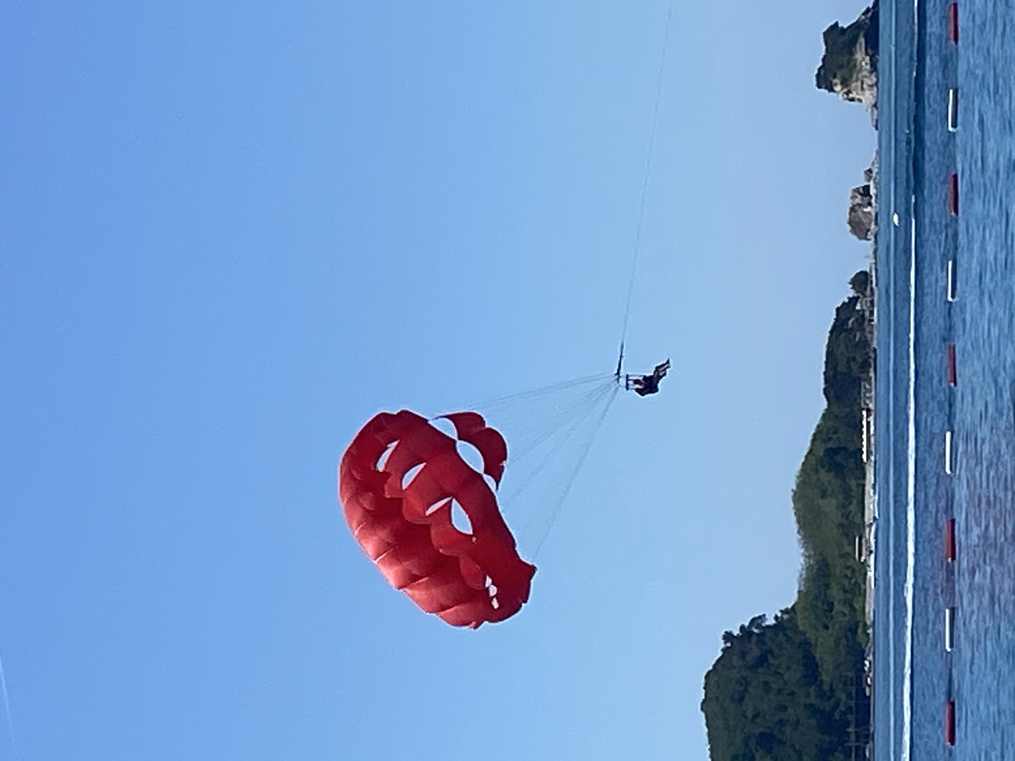 An individual is parasailing under a large, red canopy featuring multiple white, crescent-shaped cutouts on its underside, against a clear, bright blue sky. The person, rendered as a dark silhouette, is suspended in a harness and attached to a tow rope extending horizontally to the right, out of the frame. The lower third of the image depicts calm, deep blue water with faint surface ripples. In the foreground, a line of six cylindrical buoys, alternating red and white, floats on the water. The background reveals a lush, dark green, forested coastline on the left, with some distant structures at its base. Further out in the water, several rocky islets and a prominent two-peaked island formation are visible. A white motorboat is seen moving across the water in the middle ground, creating a small wake. This scene is identified as taking place near the coastline of Budva, Montenegro, with the distinct island formations characteristic of the area, possibly including Sveti Nikola Island.