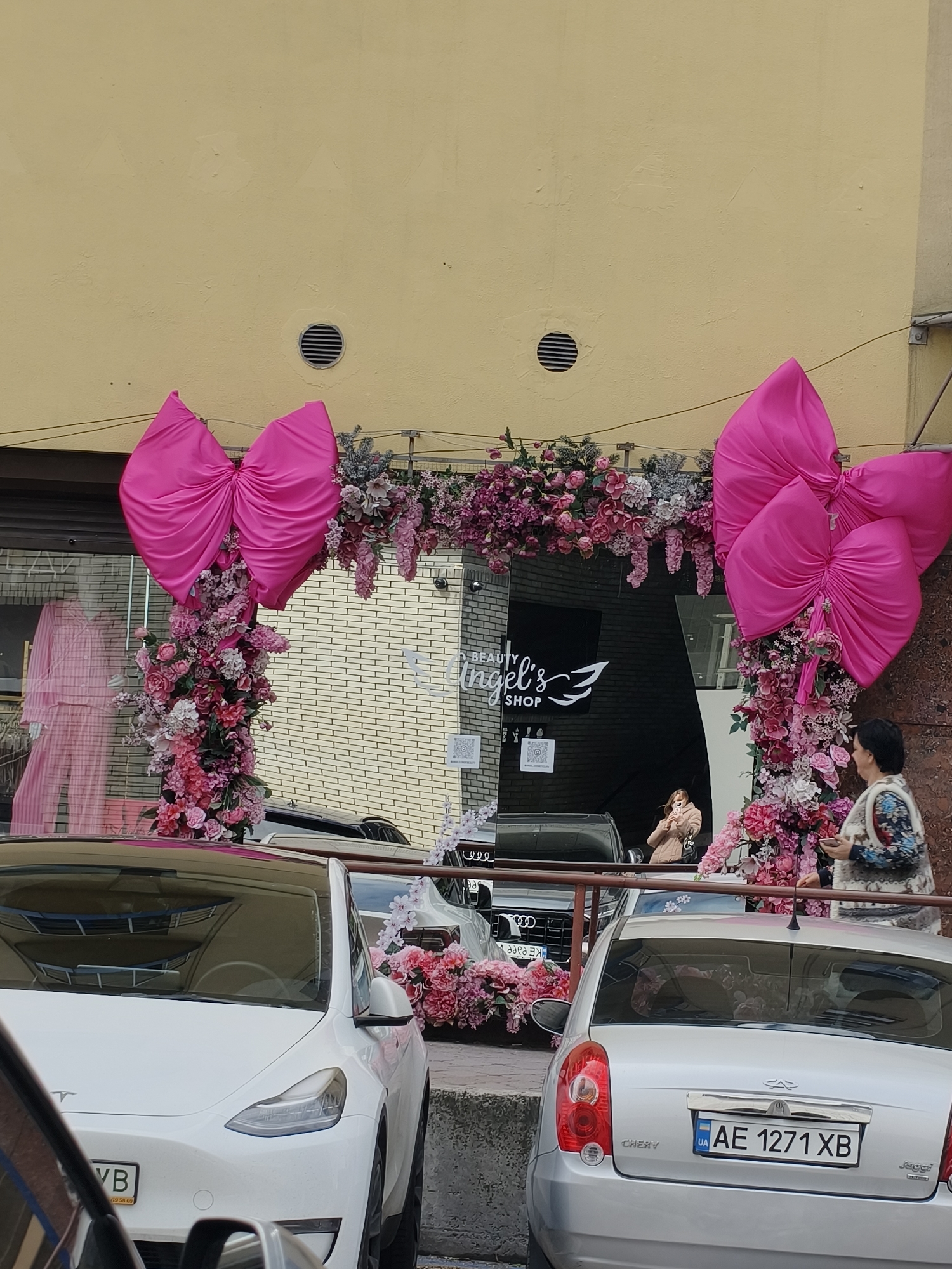 The image displays an urban street scene in Dnipro, Ukraine, centered on a shop entrance elaborately decorated with pink bows and flowers. The shop, identified by its sign as "Beauty Angel's Shop" with a winged logo, features an archway adorned with two large, vibrant pink bows and extensive artificial floral arrangements in various shades of pink, purple, and white, intermingled with green foliage.
Through the shop's glass front, a mannequin dressed in pink pajamas is visible on the left. In the reflection or inside the shop, a woman with light hair, wearing a light-colored jacket, is seen holding a phone. On the right side, partially obscured by the floral decor, another woman with dark hair, dressed in a patterned top and a light vest, stands near the shop entrance, looking towards the street or the shop's display.
Several cars are parked in the foreground. A white Tesla Model Y is on the left, with its Ukrainian license plate partially visible. A silver Chery car is parked to the right, prominently displaying its Ukrainian license plate: "AE 1271 XB". A dark Audi SUV is also visible behind the Tesla, showing its grille and logo.
The building above the shop has a plain yellow-beige facade, featuring two small circular vents. The scene occurs during the daytime, under natural light. The extensive and festive decorations suggest a special event, grand opening, or seasonal celebration for the beauty shop.