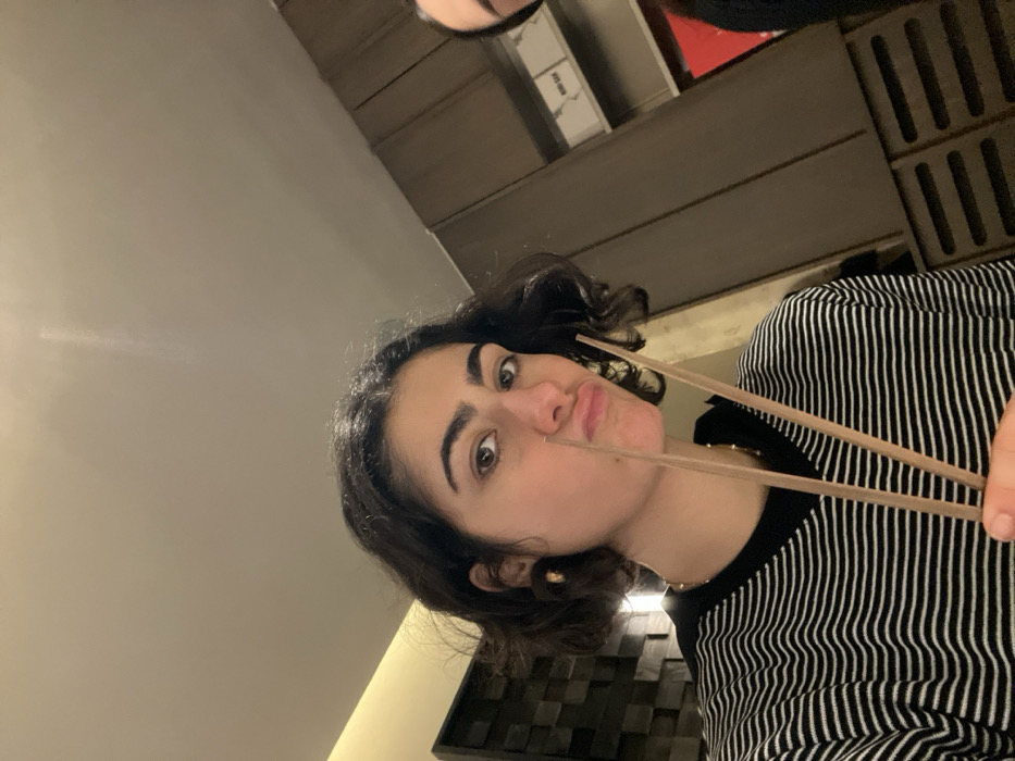 A young woman with short, dark, wavy hair holds chopsticks near her pursed lips. She has fair skin, dark eyebrows, and brown eyes. She wears a black and white striped shirt. The background includes a gray wall, a wooden paneling, and a partial view of what seems to be boxes or other stacked items. The image is rotated, with the woman's head tilted to the left.