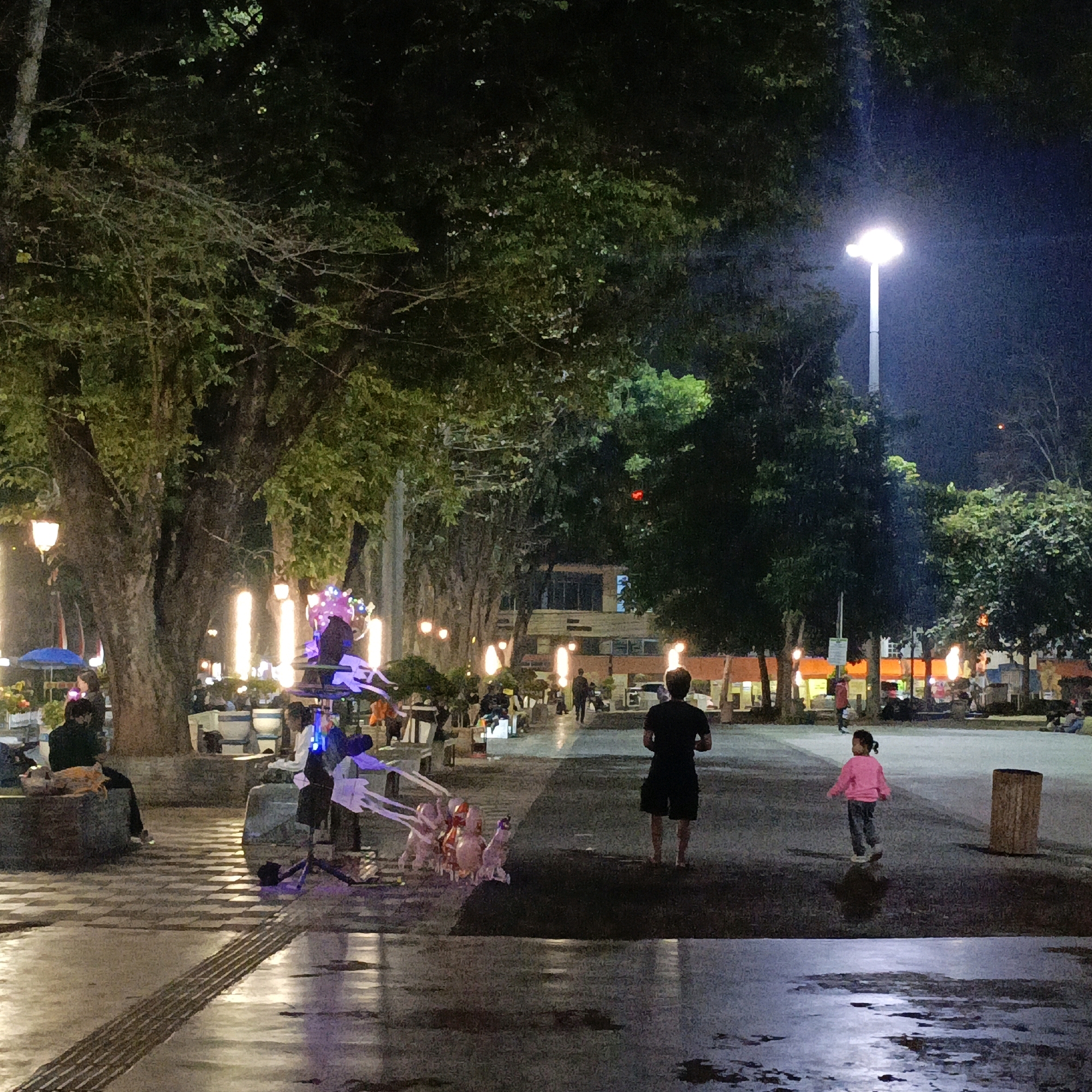 This image depicts a nighttime scene at the Pancasila Field in Salatiga, Indonesia. The field is illuminated by streetlights and decorative lights strung around trees and poles. Several people are present, including a man in a black t-shirt and shorts walking away from the camera, and a young girl in a pink jacket and jeans walking towards the right of the frame. A vendor is visible with a display of balloons. The ground appears wet in places, reflecting the ambient light. Trees line the pathway, creating a park-like atmosphere. In the background, buildings with illuminated windows suggest shops or other establishments. The overall ambiance is lively and social, characteristic of a public gathering space at night.