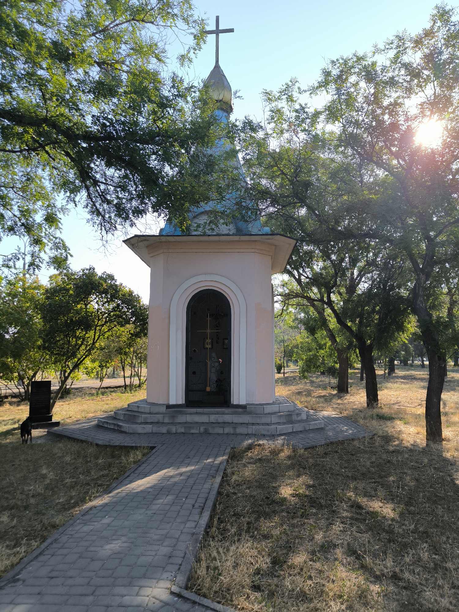 A small, pink chapel with a blue roof topped by a golden cross stands in a park-like setting. A paved walkway leads to the chapel's arched entrance, which features a dark metal gate. The chapel is surrounded by dry grass and trees, with sunlight filtering through the leaves, creating dappled shadows. In the foreground, to the left, a dark monument stands next to a small black dog. The scene appears to be a quiet, outdoor space, possibly a cemetery or memorial park. The location is the Chapel of the Holy Cross in the city of Mykolaiv, Ukraine, specifically within the cemetery grounds on Prospekt Myru.
