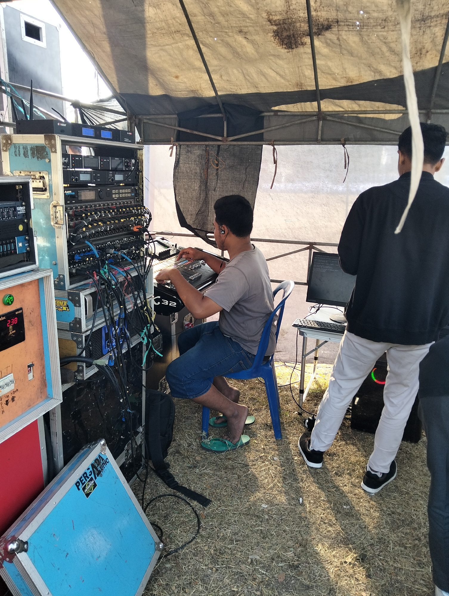 In the city of Paciran, Indonesia, under a makeshift tent, a man wearing a grey t-shirt and denim shorts is seated on a blue plastic chair, operating a sound mixing board. He is wearing earbuds and appears focused on his task. To his right, another person in a black jacket and light-colored pants stands, facing a computer monitor and keyboard on a small table. The scene is dominated by a complex setup of audio equipment, including racks of amplifiers, mixers, and numerous cables. Various cases and equipment are placed around the area, including a blue case with "PERSADA AUDIO" written on it. The ground is covered with dry grass and some debris. The overall environment suggests an outdoor event or performance setup.