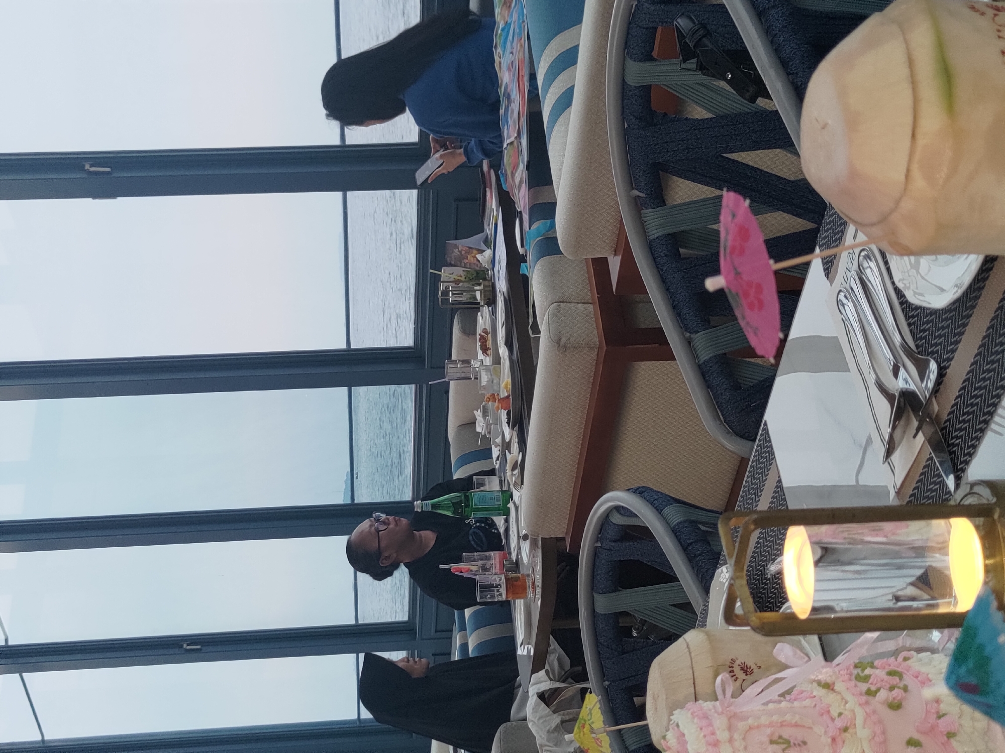 In a restaurant with a waterfront view, three women are seated at tables. The woman on the left, wearing a black hijab, is looking towards the woman in the center. The central woman, with her hair in a bun and wearing glasses, is looking to her right. To the right, a woman with long dark hair is looking down at her phone. On the tables, there are drinks, food, and a lit lantern. The restaurant has blue and white striped seating and is illuminated by natural light from large windows overlooking the water. A decorative cake and coconuts are visible on a foreground table. The location is identified as Kosambi Barat, Indonesia.