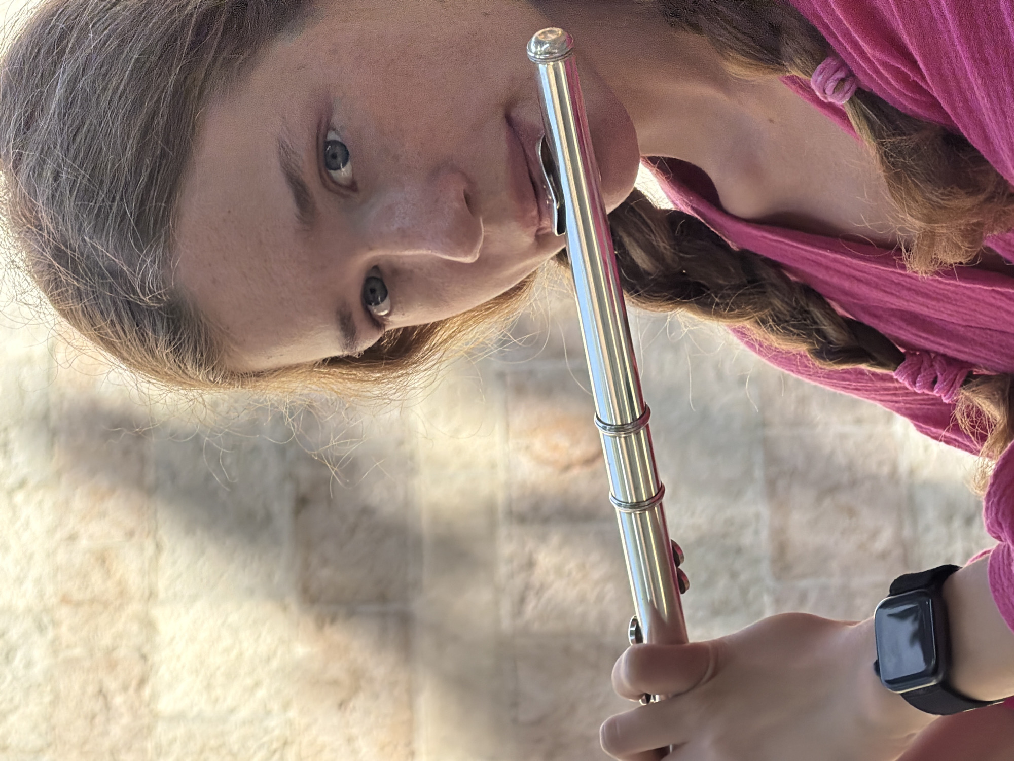 A young woman is holding a flute to her lips. She is wearing a pink top, and her hair is braided in two braids with pink ties. The background appears to be a textured wall, and the location may be the city of Budva, Montenegro. The woman is wearing a black smartwatch on her left wrist.