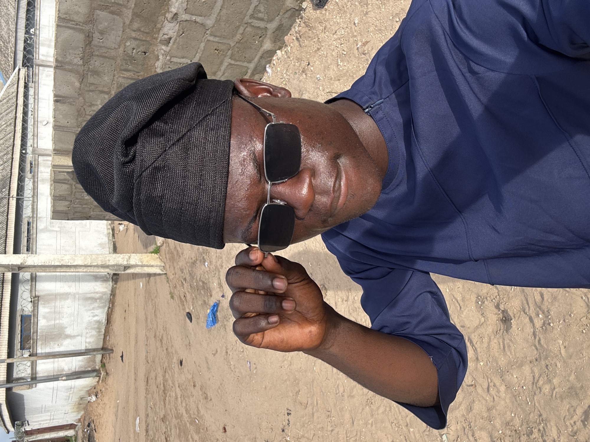 A dark-skinned person wearing a black cap, sunglasses, and a navy blue shirt is posing in a portrait setting. The person is holding their sunglasses. The location appears to be near a building with what looks like a barbed wire fence. The ground consists of sand, and there are some scattered debris, including a blue plastic item. Given the prompt, the location is speculated to be in the city of Badagry, Nigeria.