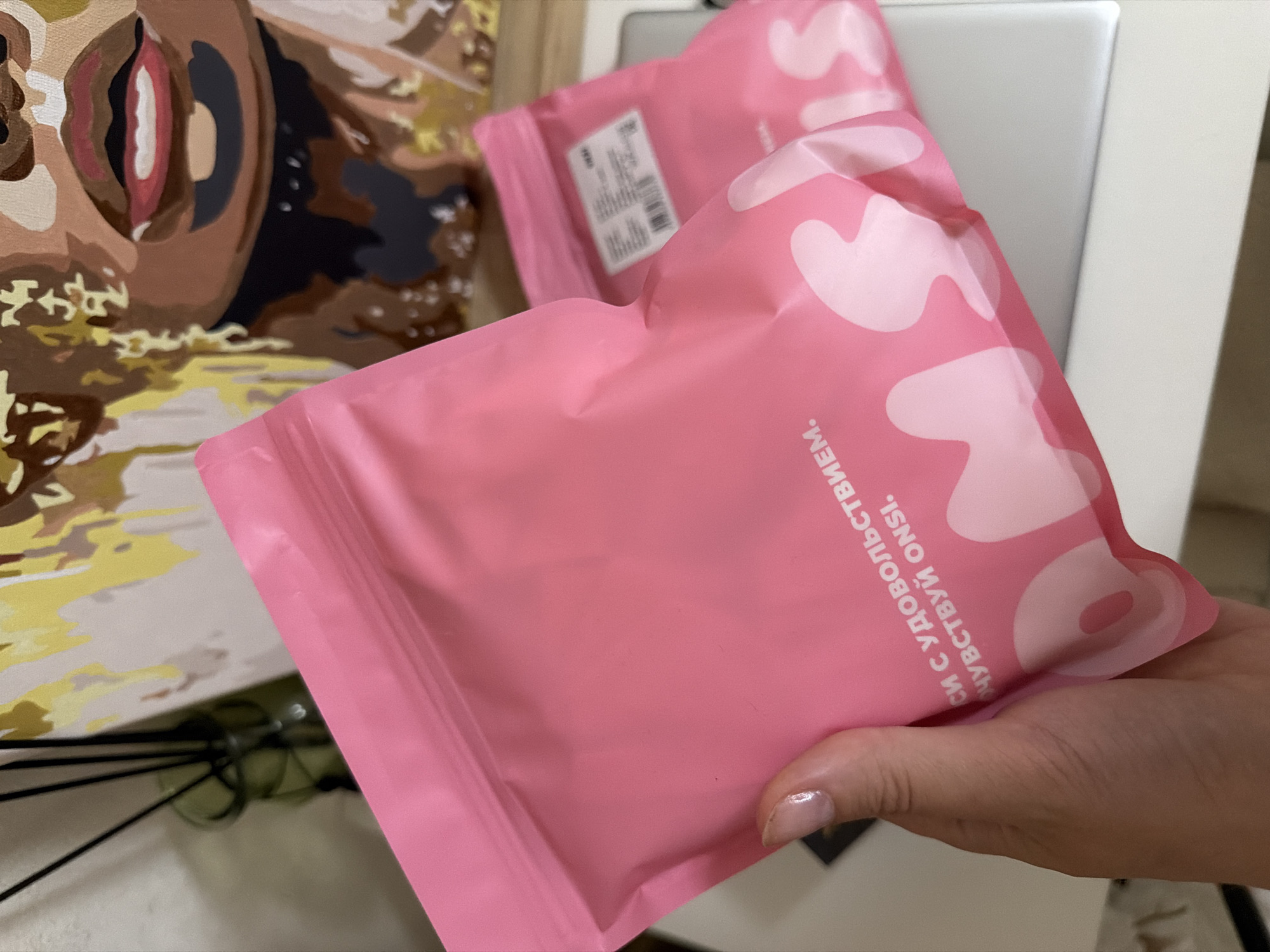 A hand holding a pink, resealable pouch with white lettering is featured in the foreground. The pouch appears to be part of a product or packaging. Behind it, another similar pouch is visible, with a white label and barcode. In the background, a partially visible painting of a person's face with dark skin and colorful makeup is displayed. The overall setting suggests an indoor environment, possibly a home or studio. The image was taken in the city of Semiluki, Russia.