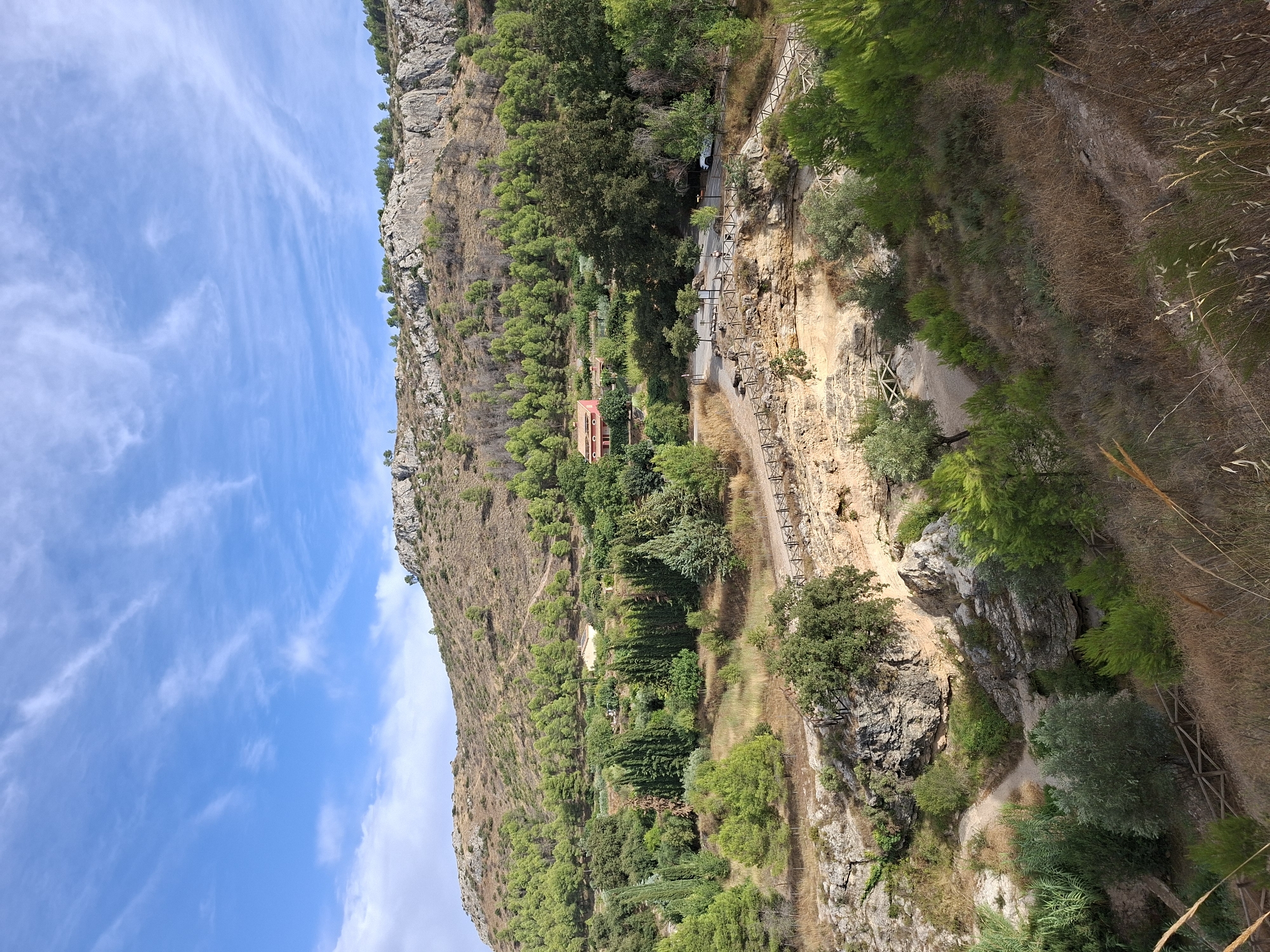 The image shows a rocky hillside with scattered trees and vegetation under a bright blue sky with wispy white clouds. A pathway, bordered by a wooden fence, winds through the terrain. In the midground, there is a cluster of trees and a red-roofed building. A white car is visible on a paved road that crosses the scene. In the foreground, the landscape is more rugged with dry grass and sparse bushes. The scene appears to be a natural landscape with some man-made elements like pathways and a building, suggesting it could be a park or a rural area. Based on the provided context, this location is identified as the Salto del Usero in Bullas, Spain.