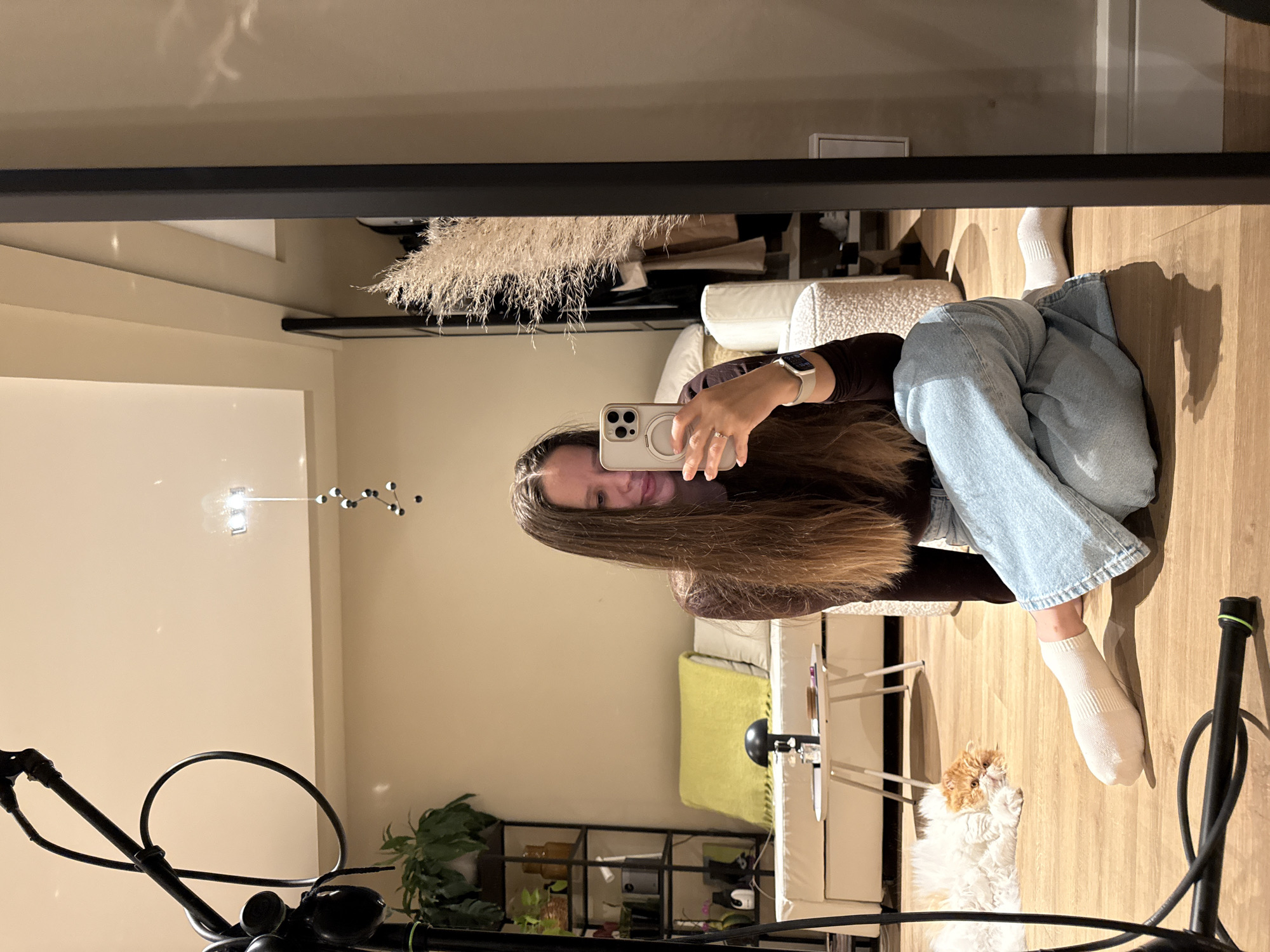 A female subject is depicted indoors, seated cross-legged on a light brown wooden floor, taking a mirror selfie in a residential setting in Barcelona, Spain. She is wearing a dark long-sleeved top, light blue jeans, and white socks. Her long, light brown hair is visible past her shoulders. She holds a light-colored smartphone (specifically an iPhone with a triple camera array and a MagSafe ring case) in her right hand, pointed towards the black-framed full-length mirror which captures her reflection. A light-colored smartwatch is on her left wrist, and a ring is visible on her left ring finger. Her expression indicates a slight smile.
To the subject's left, a fluffy orange and white cat lies resting on the wooden floor. Behind the subject, a white sofa or daybed is visible, adorned with a bright yellow throw blanket and a white pillow. A small, white, two-tiered coffee table with a dark table lamp and other small objects is positioned in front of the sofa. Further left, a tall, dark metal shelving unit displays potted plants and miscellaneous items. To the right of the sofa, a white textured armchair or stool is partially visible. A large arrangement of dried pampas grass stands behind the right side of the mirror.
In the foreground on the left, a black light stand or similar piece of equipment with cables and a green band is partially visible. The room features light-colored walls and a ceiling with architectural details, from which a modern pendant light fixture with multiple dark spheres is suspended.