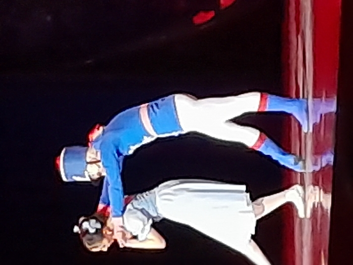 The image captures a theatrical performance featuring two individuals, likely children or young performers, executing a synchronized pose on a stage. The image is oriented vertically, depicting the scene as if viewed from the side with the performers appearing to defy gravity, but it represents a floor-based act on a reflective surface.
The upper figure, when viewed with correct orientation, is a performer dressed in a costume resembling a toy soldier or a military uniform. This costume includes a blue jacket with red and white accents, white trousers with red bands at the knees and blue lower legs/boots, and a blue hat with a red band and a white plume. This individual is in an arched, bridge-like pose, with their body bent backwards. Their hands are positioned near the second performer, while their legs are extended back.
The lower figure is lying on their back beneath the first performer. This individual wears a light-colored, possibly white or light blue, dress with short sleeves and a full skirt. Their dark hair is tied back and adorned with a white bow or hair accessory. Their pose suggests they are supporting or interacting with the arched figure, with hands positioned near their head or the first performer's hands. One leg is slightly bent.
The background is predominantly dark, characteristic of a stage setting, which emphasizes the brightly lit performers. On the right side of the frame, there is a vertical element with red and white stripes, which could be a stage prop, a curtain, or part of the set design. A notable detail is the highly reflective stage floor, which creates clear, almost mirror-like reflections of both performers and the striped vertical element beneath them. The scene suggests a ballet, dance, or acrobatic act, possibly from a production with a toy soldier theme, such as The Nutcracker. The performers appear focused and skilled in their coordinated movement. No text is visible in the image.