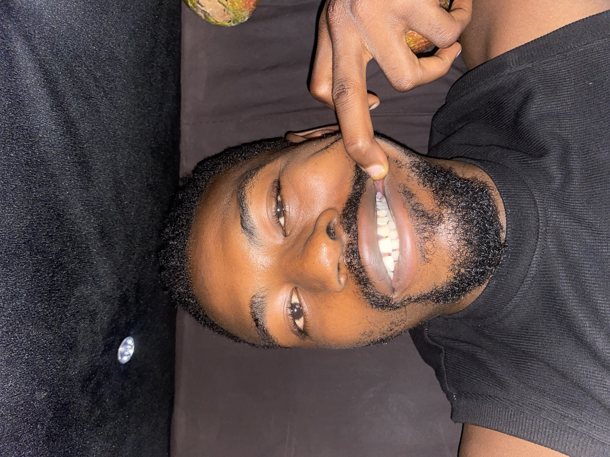A young man with dark skin and a short, dark beard is depicted lying on his back on a dark, textured surface, likely a couch or bed. He is wearing a black, ribbed t-shirt. His head is slightly tilted, and he is looking directly at the camera with open eyes. His left index finger is playfully pulling up his upper lip, revealing his bright white teeth and gums. His expression conveys amusement or playfulness. The lighting appears to be artificial, suggesting an indoor setting in Barakin Akawo, Nigeria. In the top left corner of the frame, a small portion of a green and brown textured object, possibly a fruit, is visible. A small, reflective object can also be seen on the dark surface near the bottom left. There is no visible text in the image.