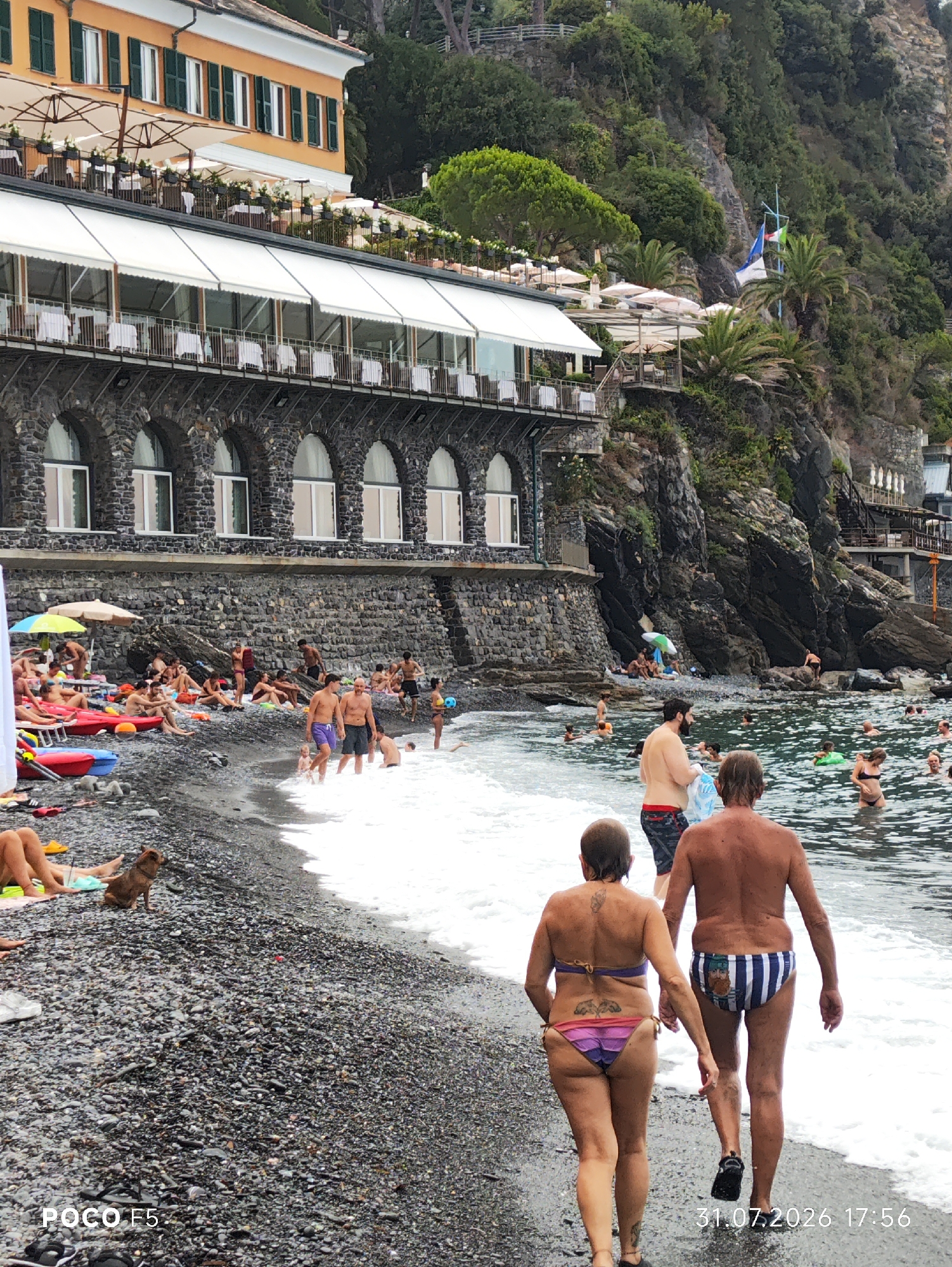The image depicts a beach scene in Camogli, Italy, with a focus on a pebble beach and the Ligurian Sea. In the foreground, a couple walks away from the camera towards the water. The woman is wearing a purple and pink bikini, and the man is wearing striped swim trunks. Behind them, the sea is filled with people swimming and wading. Some are using inflatable rings, and one person is playing with a yellow ball.
Further back on the beach, numerous people are sunbathing on towels and loungers. Red and yellow inflatable kayaks are visible near the water's edge. A small brown dog is sitting on the pebbles in the lower left portion of the frame.
Dominating the background is a multi-story building with an orange facade, featuring dark green shutters and awnings extending over a lower level with arched stone openings. This building appears to be a hotel or restaurant, with tables and chairs set up on an outdoor terrace. Beyond the building, a steep, rocky hillside is covered in lush green vegetation. Flags, including the Italian tricolor, are visible on a pole on the hillside. The overall atmosphere is one of a busy summer day at the beach. The date stamp "31.07.2026 17:56" and "POCO F5" are present in the lower corners of the image.