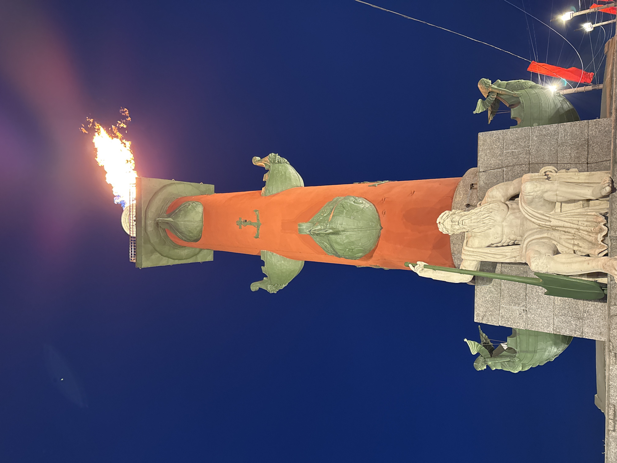 The image shows the Rostral Columns in St. Petersburg, Russia, illuminated at night. The orange column is adorned with green rostral prows, and atop one column, a flame burns brightly. A sculpture of a reclining figure is visible at the base of the column, as are additional sculptures on the pedestal. The background is a dark blue night sky. Some hanging lights and wires are visible in the upper right.