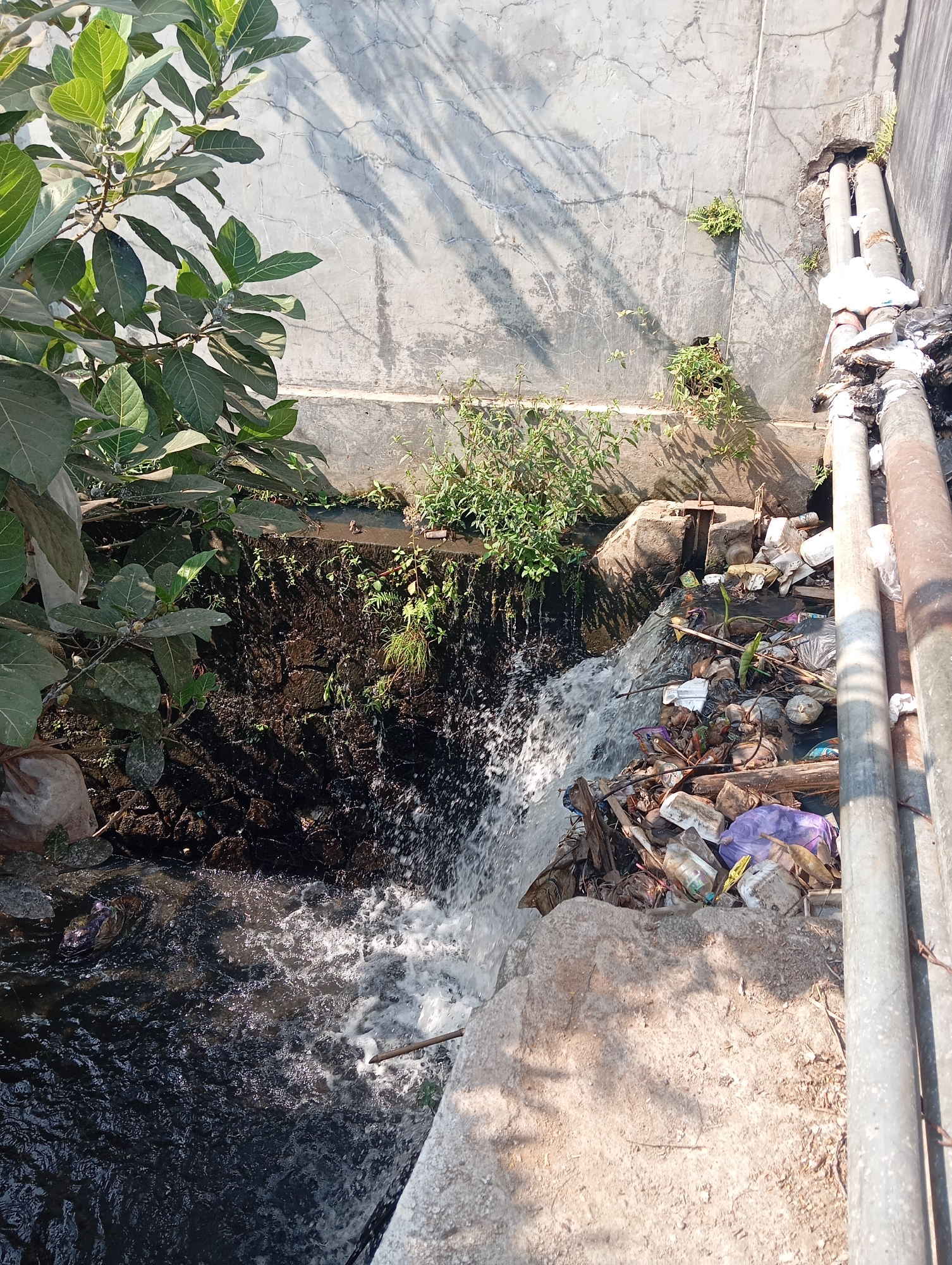 The image depicts a polluted drainage channel with a small waterfall. The channel is filled with various types of debris, including plastic bottles, bags, wood, and other refuse. The water is murky and appears to be flowing through the trash. Green foliage grows along the sides of the channel and on the concrete wall behind it. Two large metal pipes run vertically along the right side of the image, and a concrete structure is visible to the left. The scene suggests a lack of proper waste management and environmental upkeep, possibly in an urban or semi-urban area like Sokaraja, Indonesia.