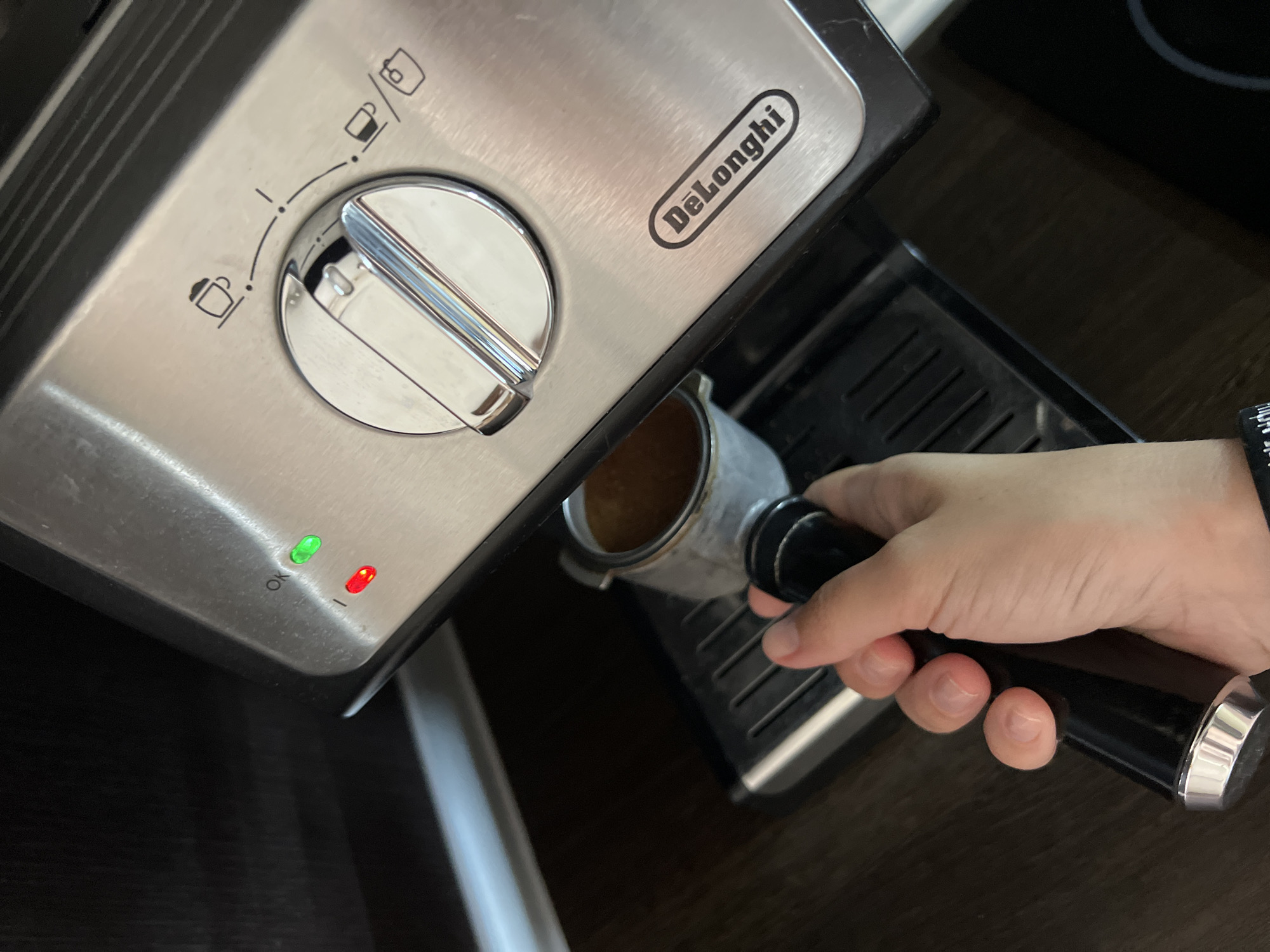 In an indoor setting, a person's hand is holding the handle of an espresso machine's portafilter, which is inserted into the machine. The espresso machine is made of brushed stainless steel and has a dial with icons indicating different brewing options. There are two indicator lights on the machine, one green and one red, with the green light illuminated. The portafilter contains ground coffee. The background shows a dark countertop and cabinetry.