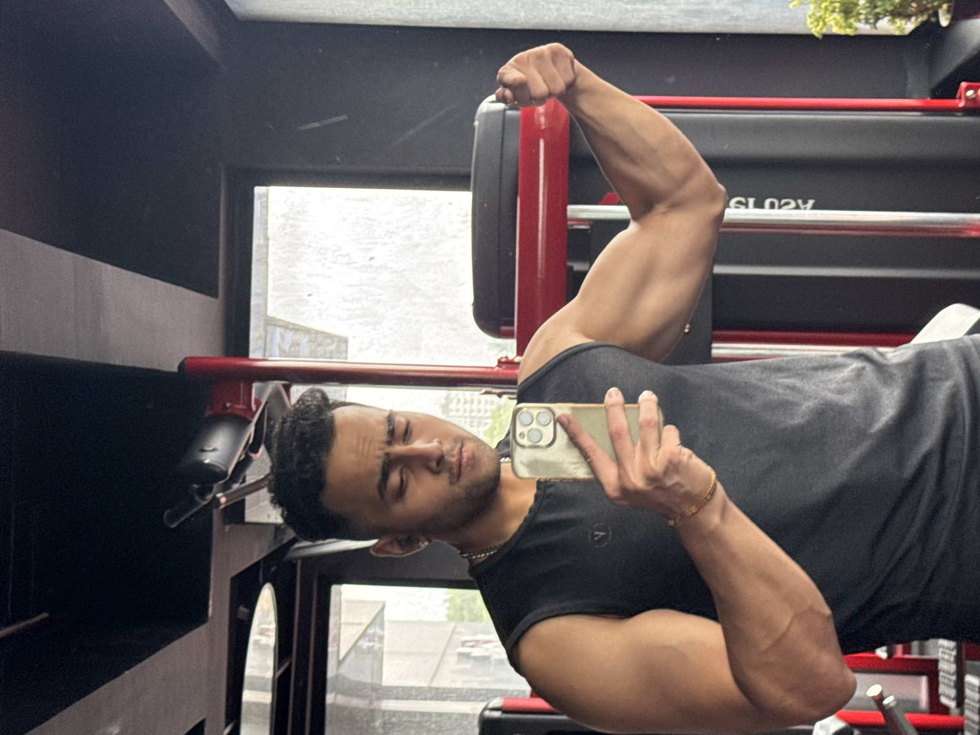 The image is a selfie taken in a gym. A young man with short, dark hair and a light beard is looking at his phone, which he is holding in his left hand. He is wearing a dark grey, sleeveless t-shirt and a gold-colored chain around his neck. His right arm is flexed, showing his bicep. The gym equipment is visible in the background, including red and black metal structures and a weight plate. The environment appears to be an indoor gym with natural light coming from windows. The city of Kolkata, India is not discernible from the provided image.