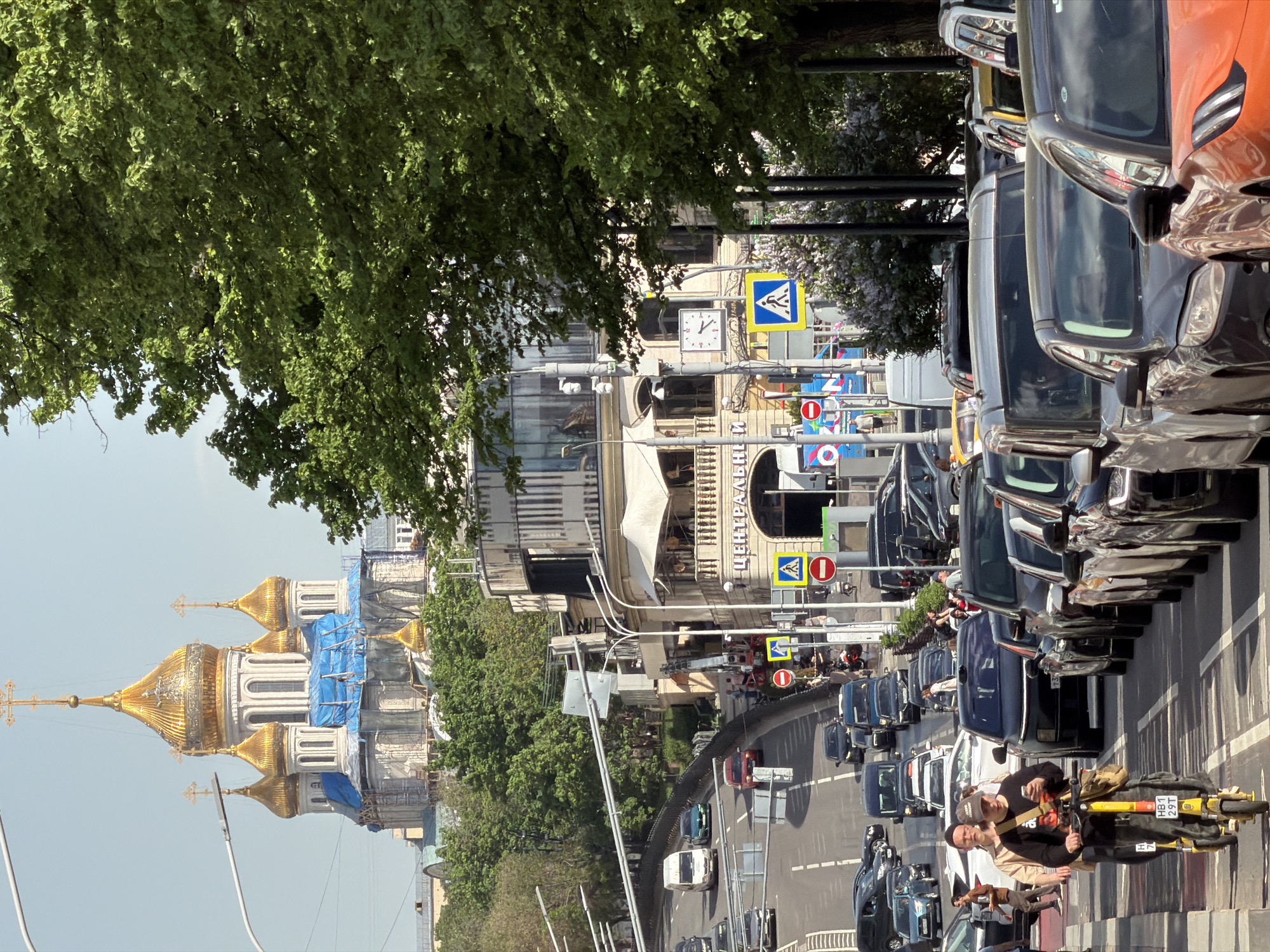A street scene in Moscow, Russia, is depicted in a high-angle, full shot taken on a sunny day. On the left, an ornate Russian Orthodox church with multiple golden domes under partial blue tarp renovation rises in the background. A dense green tree canopy extends across the upper left portion of the image, partially obscuring a mix of building styles. The street is filled with cars parked in a long line on the right, and more cars are seen driving down the street. Pedestrians are visible walking near the parked cars, and two men are standing with a yellow scooter on the sidewalk. Pedestrian crossing signs and other street signs are also visible.