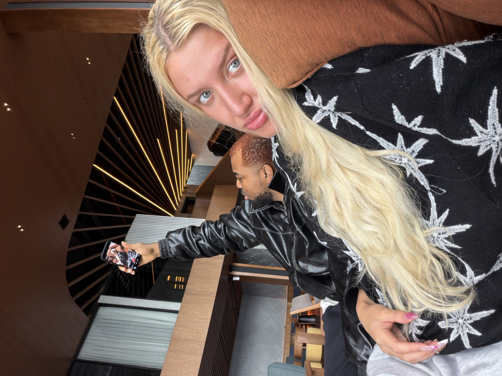 In a dimly lit indoor space with wooden architectural elements and modern lighting, two individuals are captured in a close-up selfie. On the right, a person with long blonde hair and blue eyes looks directly at the camera, resting their head on an orange cushion. They are wearing a black garment with a white star pattern. To the left and slightly behind, another person with short, reddish-brown hair and a beard is seen from the shoulders up, wearing a black leather jacket. This individual is holding up a smartphone, seemingly taking the selfie, and their face is partially obscured. The background reveals a contemporary interior with visible structural beams and lighting fixtures, suggesting a restaurant or lounge area. The location is Kintamani, Indonesia.