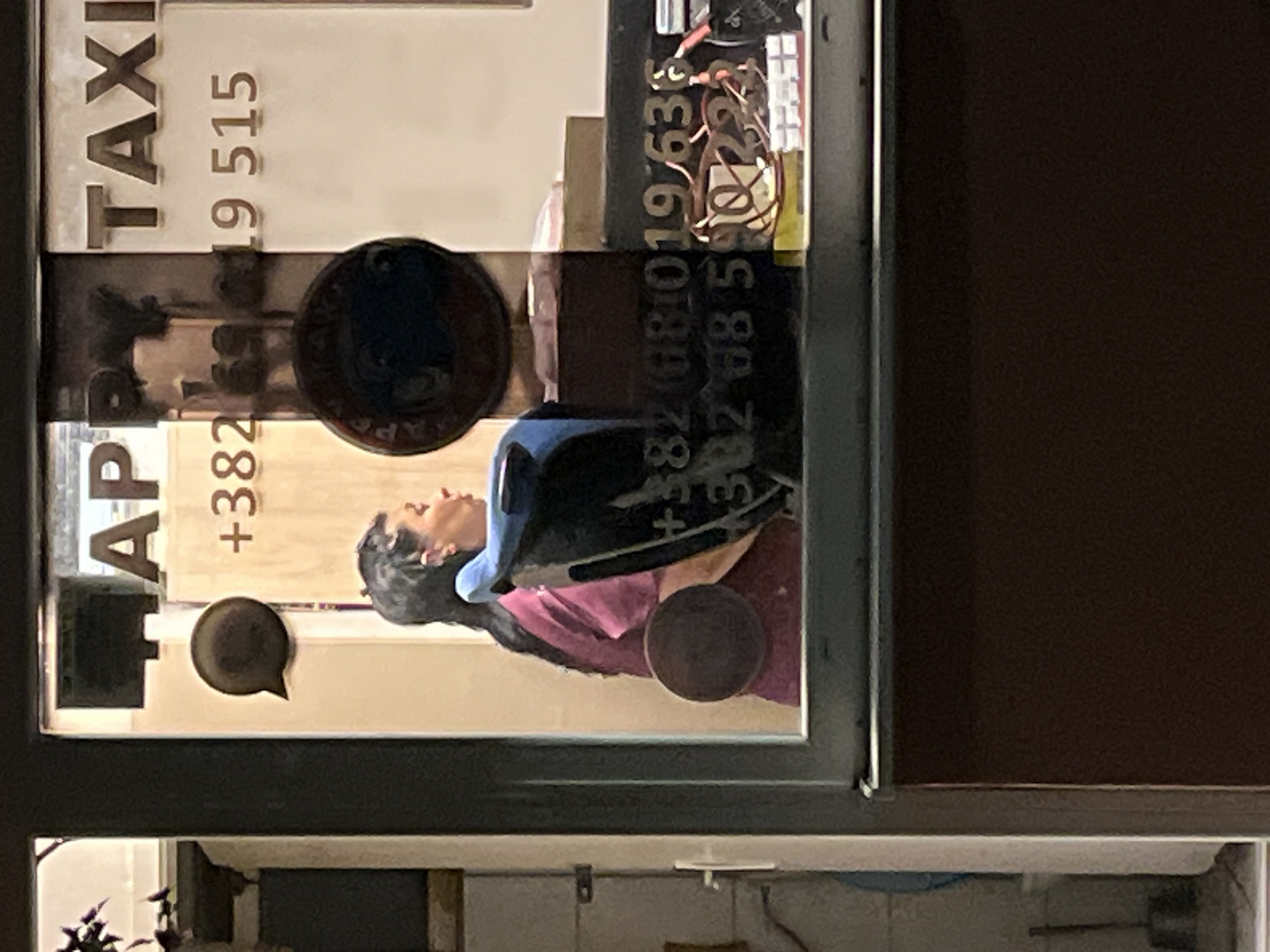 The image shows the interior of a taxi dispatch office with a woman visible behind a counter. The text on the window reads "HAPPY TAXI" and displays two phone numbers: "+382 69 619 515" and "+382 68 590 222". The office appears to be located in Budva, Montenegro. The woman is looking away from the camera and appears to be working at a computer. There are several objects on the counter, including a headset and some paperwork. The lighting is dim, suggesting it is nighttime.