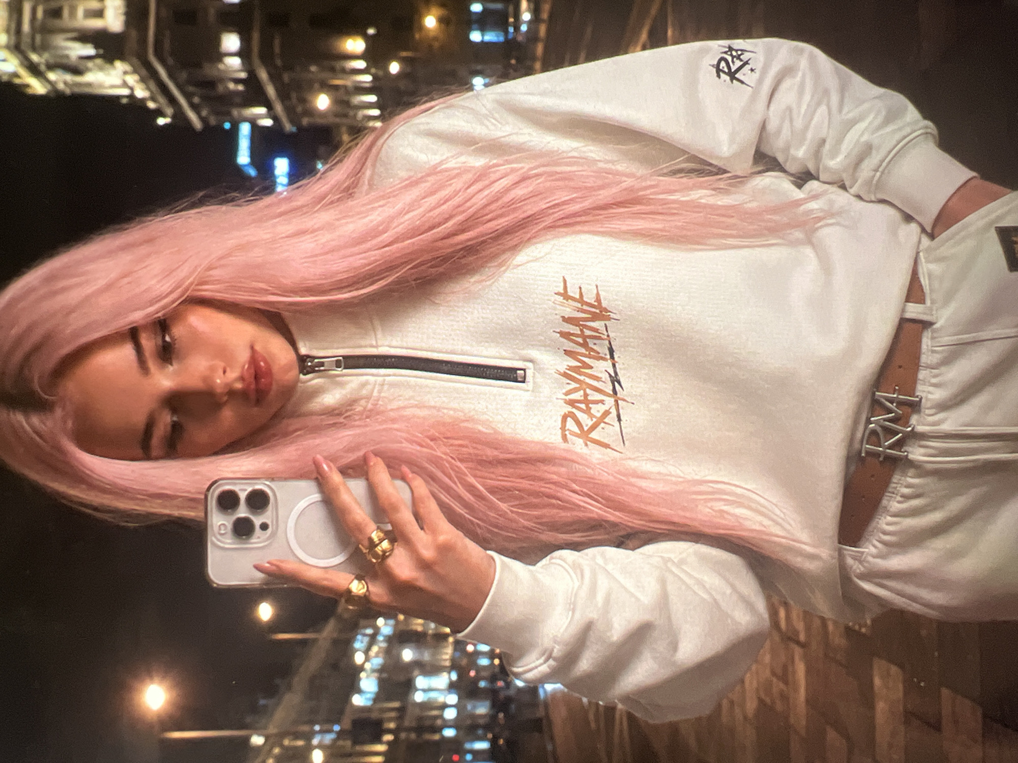 A young woman with long, pink hair takes a selfie in the city of Minsk, Belarus. She is wearing a white quarter-zip sweatshirt with orange and black text across the chest that reads "RAYMANE," and white sweatpants. She has a brown belt with a silver "RM" buckle. The background shows a nighttime street scene with illuminated buildings and streetlights.