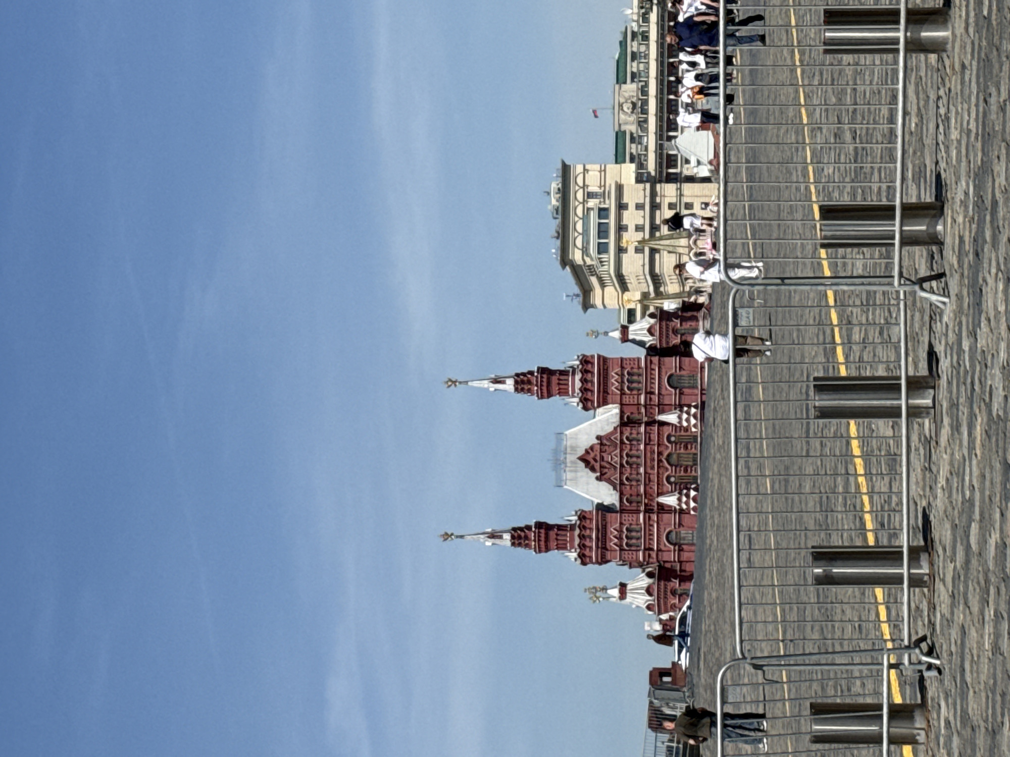 The image depicts a view of the Moscow Kremlin in Moscow, Russia, under a clear blue sky. The iconic red brick towers of the Kremlin are prominently visible, with their distinctive spires. A section of Red Square is visible to the right, with a grey stone surface and yellow lane markings. Metal crowd control barriers are set up along the square. The facades of buildings, including the GUM department store, are seen in the background. People can be seen walking and standing along the square.