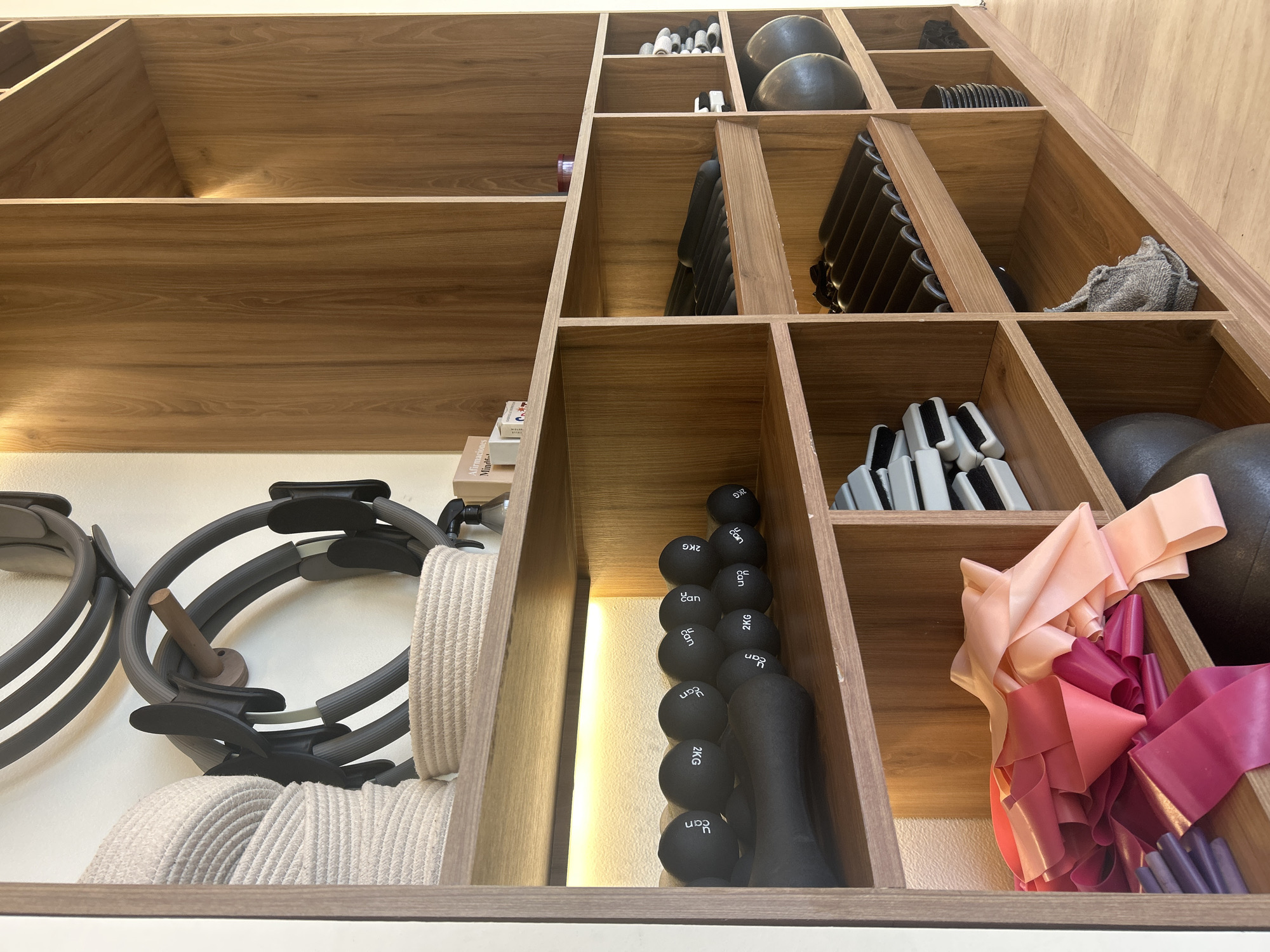 A low-angle shot depicts a wooden storage unit filled with various fitness equipment. The unit has multiple compartments of varying sizes. The equipment includes Pilates rings, small black weights marked "2 KG", light-colored fitness bands, gray hand weights, larger dark medicine balls, and other unidentified items. The storage unit appears to be well-organized and contains items used for exercise or rehabilitation. The location is unknown.