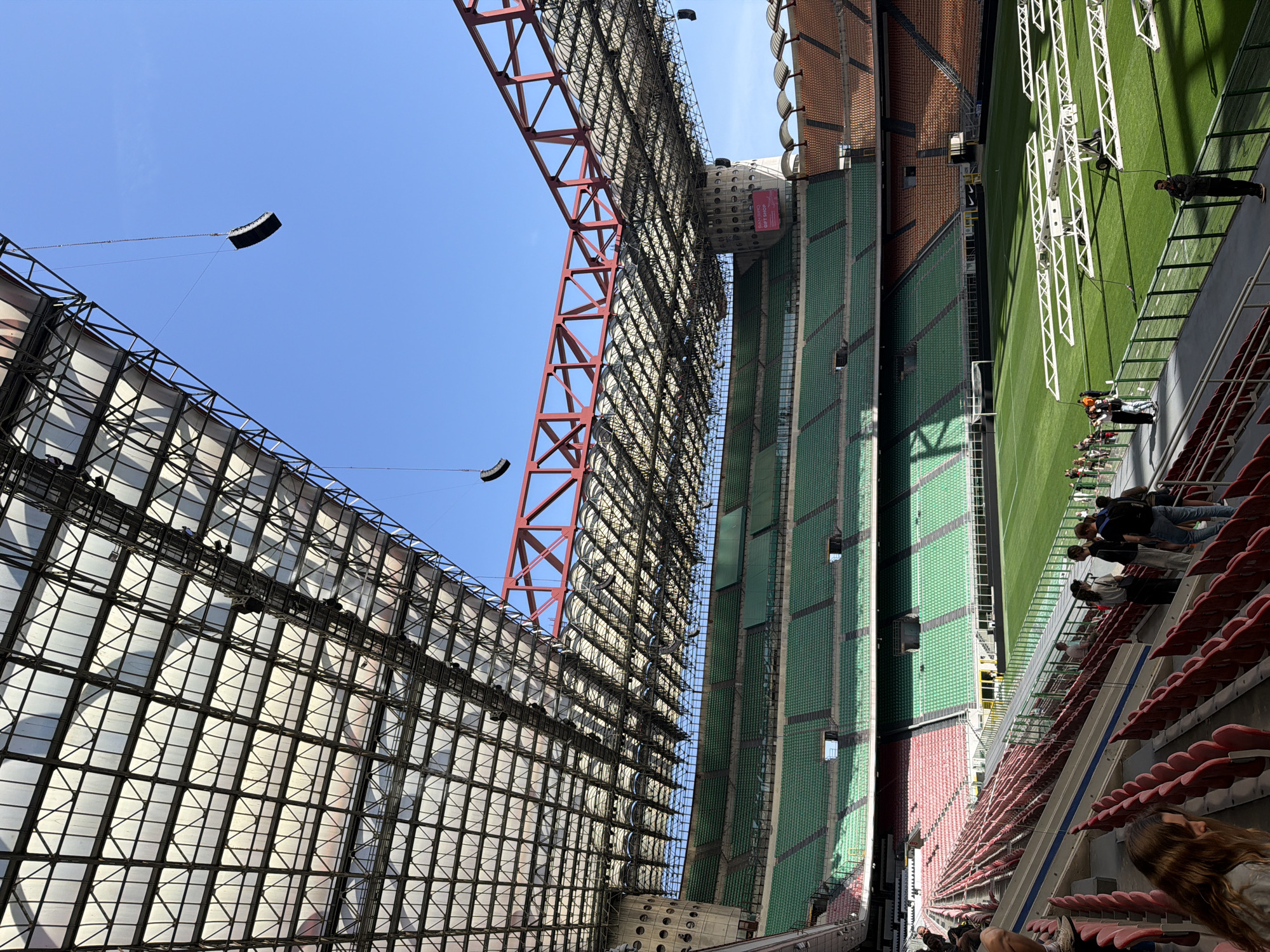 This image shows the San Siro stadium in Milan, Italy, during what appears to be preparations for an event. A large scaffolding structure and a red crane are visible on the left side of the frame, reaching towards the sky. Two rectangular black objects are suspended in the air, likely part of the equipment being installed. The seating area is visible on the right, with a mix of red and green seats. Some people are seated in the stands, while others appear to be working on the field. The environment is bright, indicating a sunny day. The overall impression is one of a stadium being prepared for an event or undergoing construction.