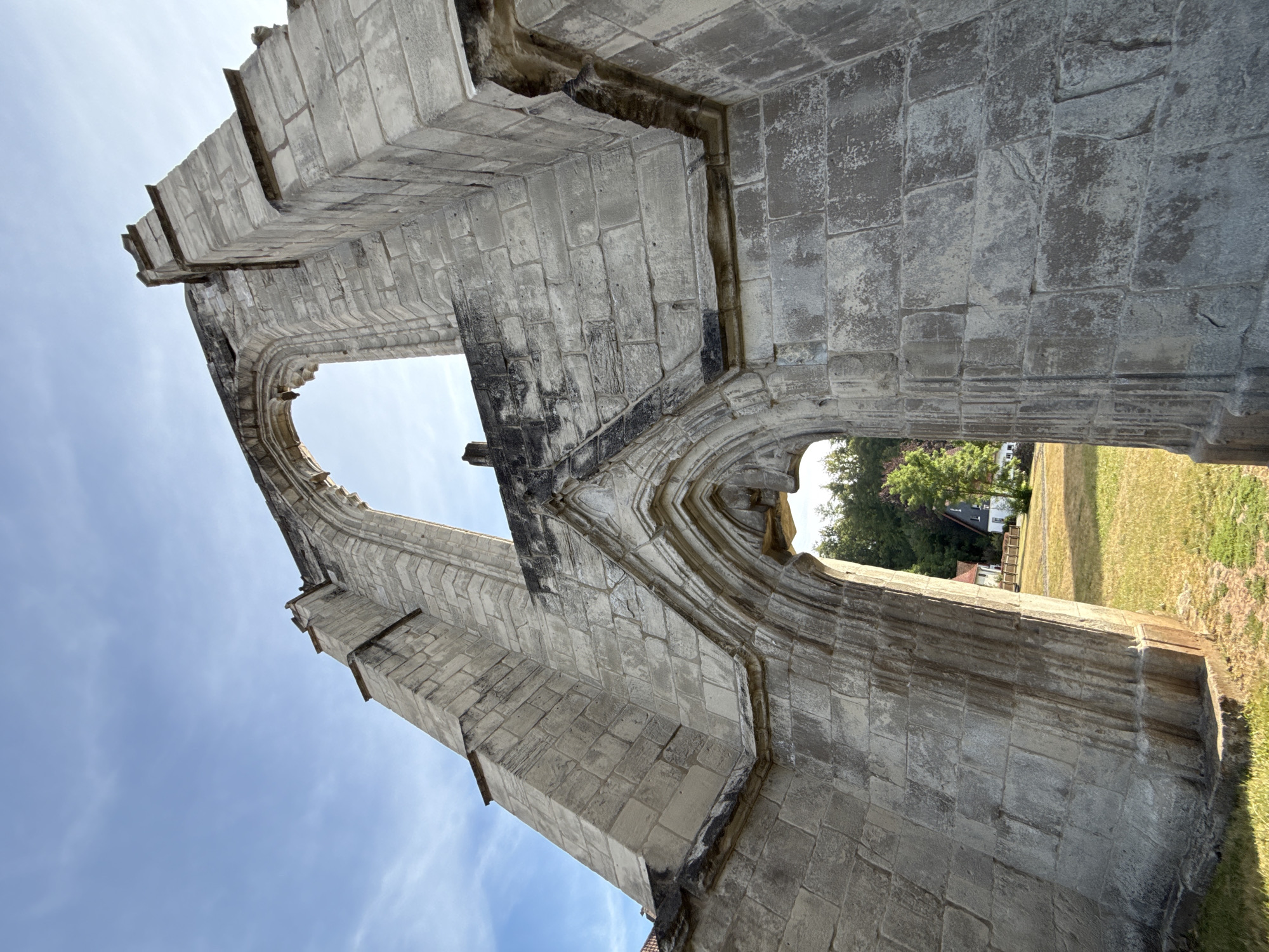 The image shows the ruin of a Gothic church in Walkenried, Germany. The structure is made of grey stone blocks and features large arched windows. In the background, a house and trees are visible. The sky is blue with some white clouds. The lighting suggests it is daytime, likely late afternoon due to the angle of the sun.