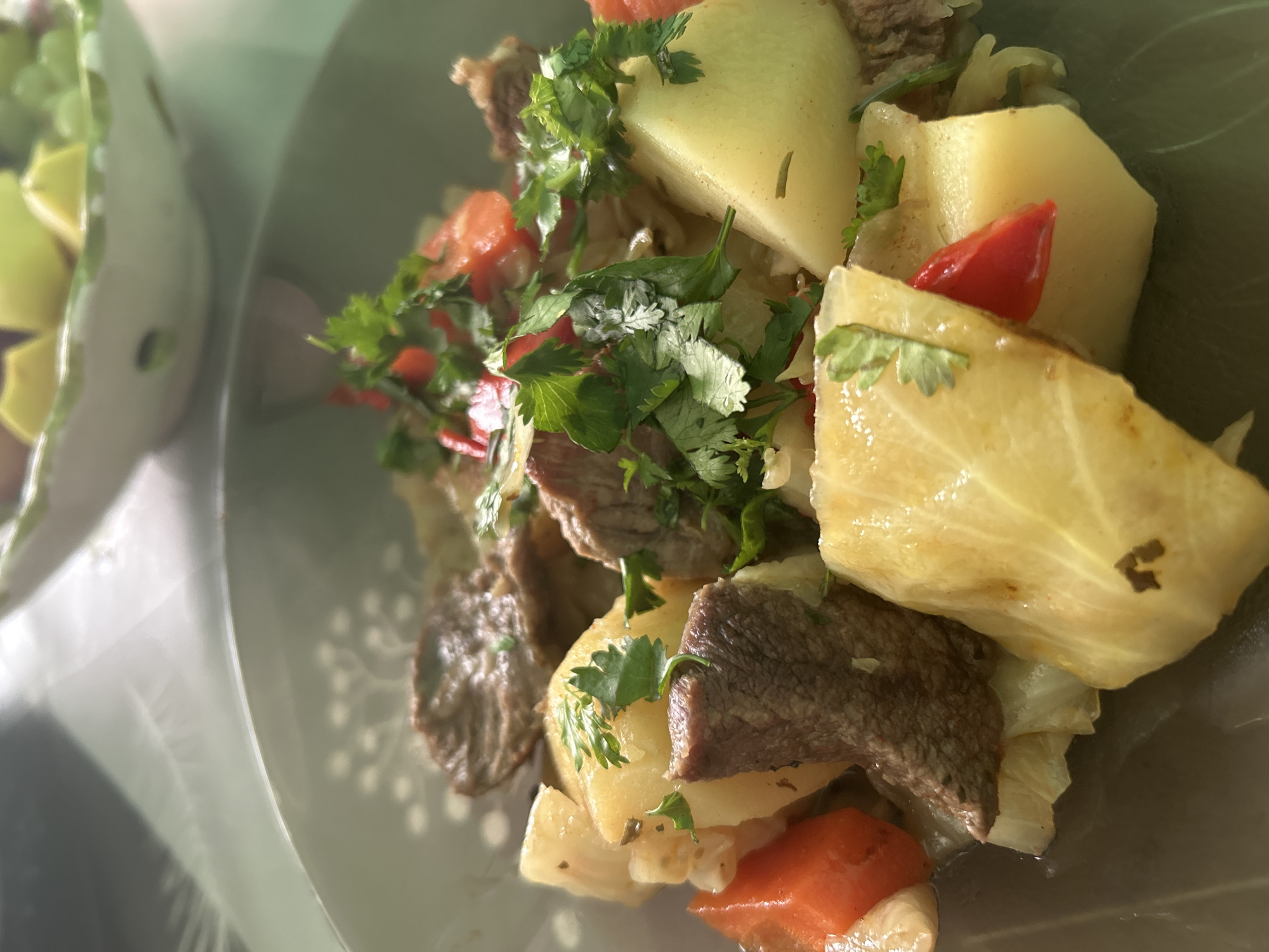 A close-up, slightly blurred, high-angle shot depicts a partially filled light green bowl containing a food dish. The main components are chunks of cooked meat, cubed potatoes, chopped red tomatoes, and leafy green herbs, likely cilantro or parsley. The dish is served on a white surface. The bowl also contains cooked cabbage. A second bowl containing sliced yellow fruits is present in the far background of the scene. The location in the city of Марковский, Russia is unconfirmed, as location data is unavailable in the image.