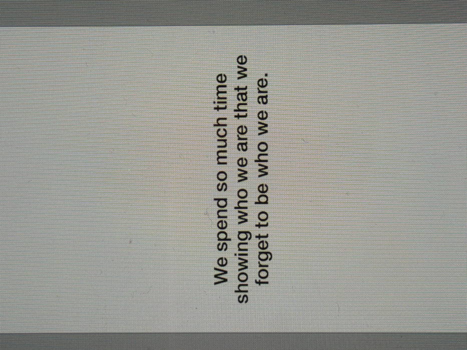 The image displays a close-up of a digital screen featuring a philosophical quote. The screen exhibits a fine, uniform vertical pixel grid across its surface, characteristic of an LCD or similar digital display. The background of the display is a light gray, and the text is rendered in a dark gray font. The quote reads: "We spend so much time showing who we are that we forget to be who we are." Darker gray vertical bands are visible along the far left and right edges of the captured frame, suggesting either the physical bezels of the screen or the extreme edges of the illuminated display area. No individuals, other subjects, objects beyond the screen, actions, or interactions are visible. The specific location within the city of London, United Kingdom, cannot be determined from the content of this image.