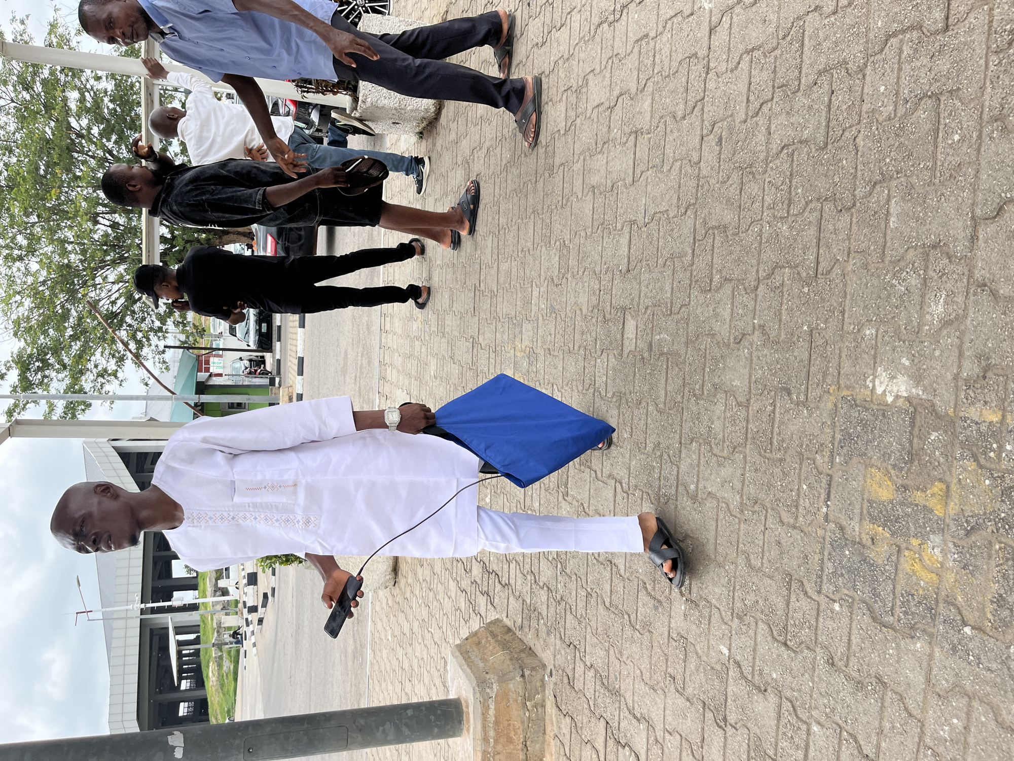 The image captures an outdoor scene in Gwaska, Nigeria, featuring several men on a paved walkway during what appears to be an overcast daytime.
In the foreground, a man wearing a white long-sleeved tunic with subtle embroidery and matching white trousers stands, holding a black smartphone in his left hand and a blue bag or folder in his right. He wears black slide sandals and a watch. His head is bald, and he is looking to his right.
Behind him and to the left, a group of other men are visible. One man in a light blue collared shirt and dark trousers stands near a pole, while another in a black short-sleeved shirt and black shorts holds a dark hat. Further back, another man in a white shirt and jeans is partially visible, and a man in a black shirt and black trousers, wearing a dark baseball cap, is also present. Most of the men are wearing sandals or slippers.
The setting is an urban or semi-urban environment with a patterned interlocking brick pavement. Green foliage from trees is visible on the left side of the frame, and modern buildings with large windows and gray/white facades are in the background. Partially visible cars are parked in the midground. The men appear to be standing around, possibly conversing or waiting. There is some unreadable text on a distant sign.