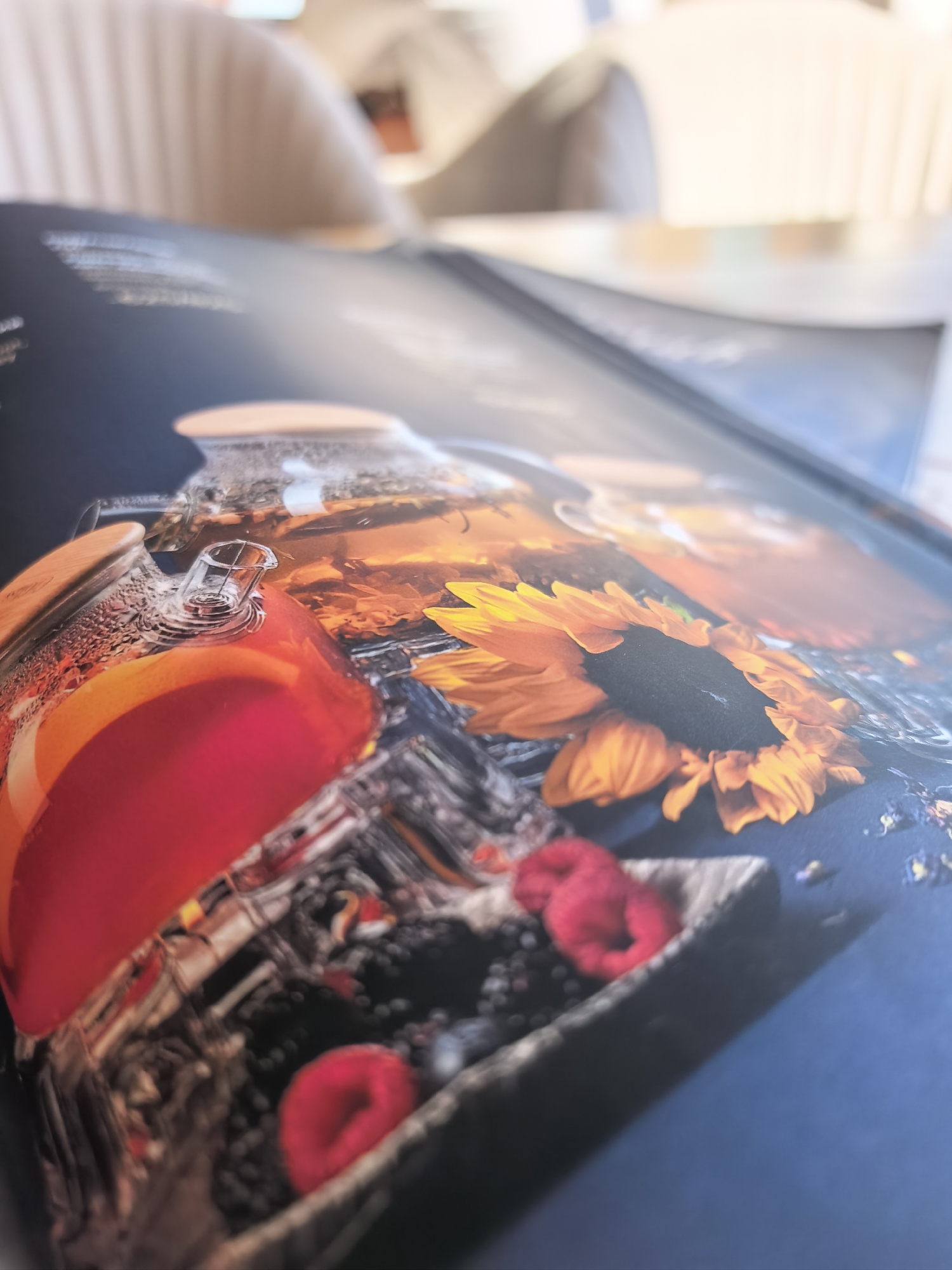 The image is a close-up, angled shot of a printed page, possibly a menu or a magazine, displaying a still life. The dominant elements are a glass teapot filled with a reddish-orange liquid and slices of citrus fruit, likely oranges. The teapot is steamy, with condensation visible on its surface. Beside the teapot, a small wooden bowl holds dark berries, possibly blackberries or blueberries, and two vibrant red raspberries. In the background of the printed image, a sunflower with bright orange petals and a dark center is visible, along with scattered dried flower petals. The page itself has a dark blue background. The actual environment appears to be a cafe or restaurant setting, with out-of-focus, pale-colored chairs in the background, suggesting a bright, ambient light. There are no people or animals present in the scene. The location in the city of Repino, Russia is not evident from the image itself.
