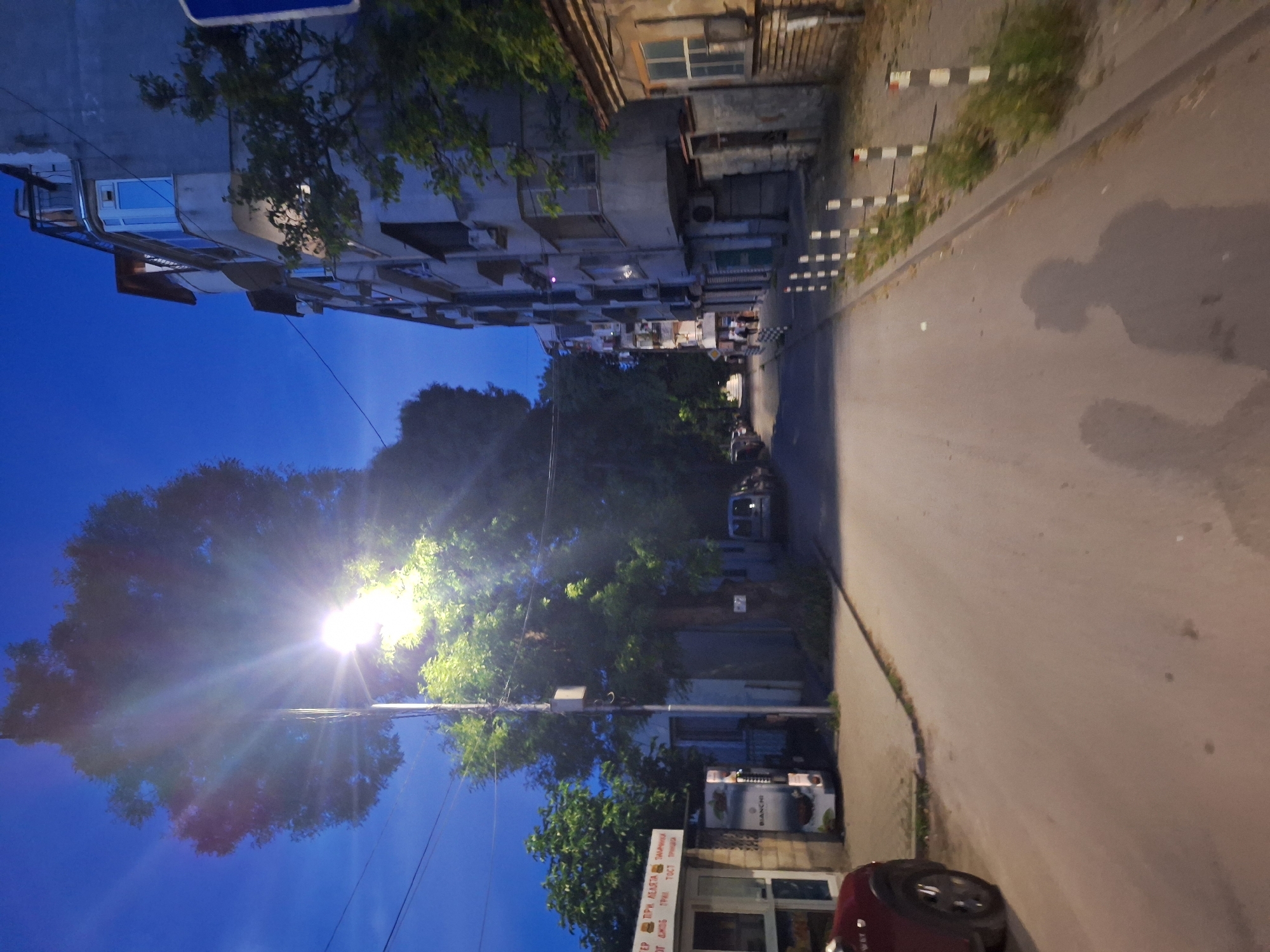 The image depicts a street scene at dusk in Varna, Bulgaria. A bright streetlight illuminates a large tree in the foreground, casting rays of light across the sky and casting shadows on the road. To the right, a multi-story residential building with several balconies and windows is visible. The building appears to be made of concrete and has a weathered facade. A narrow sidewalk runs alongside the building, lined with white and black bollards. On the left side of the street, there is a small storefront with a glass door and a sign that has some text in Cyrillic. A vending machine is also present outside the storefront. The street itself is paved and appears to be relatively empty, with a couple of cars parked along the side. Two indistinct human shadows are cast on the road in the foreground, suggesting the photographer and possibly another person are present. The sky is a deep blue, indicating twilight.