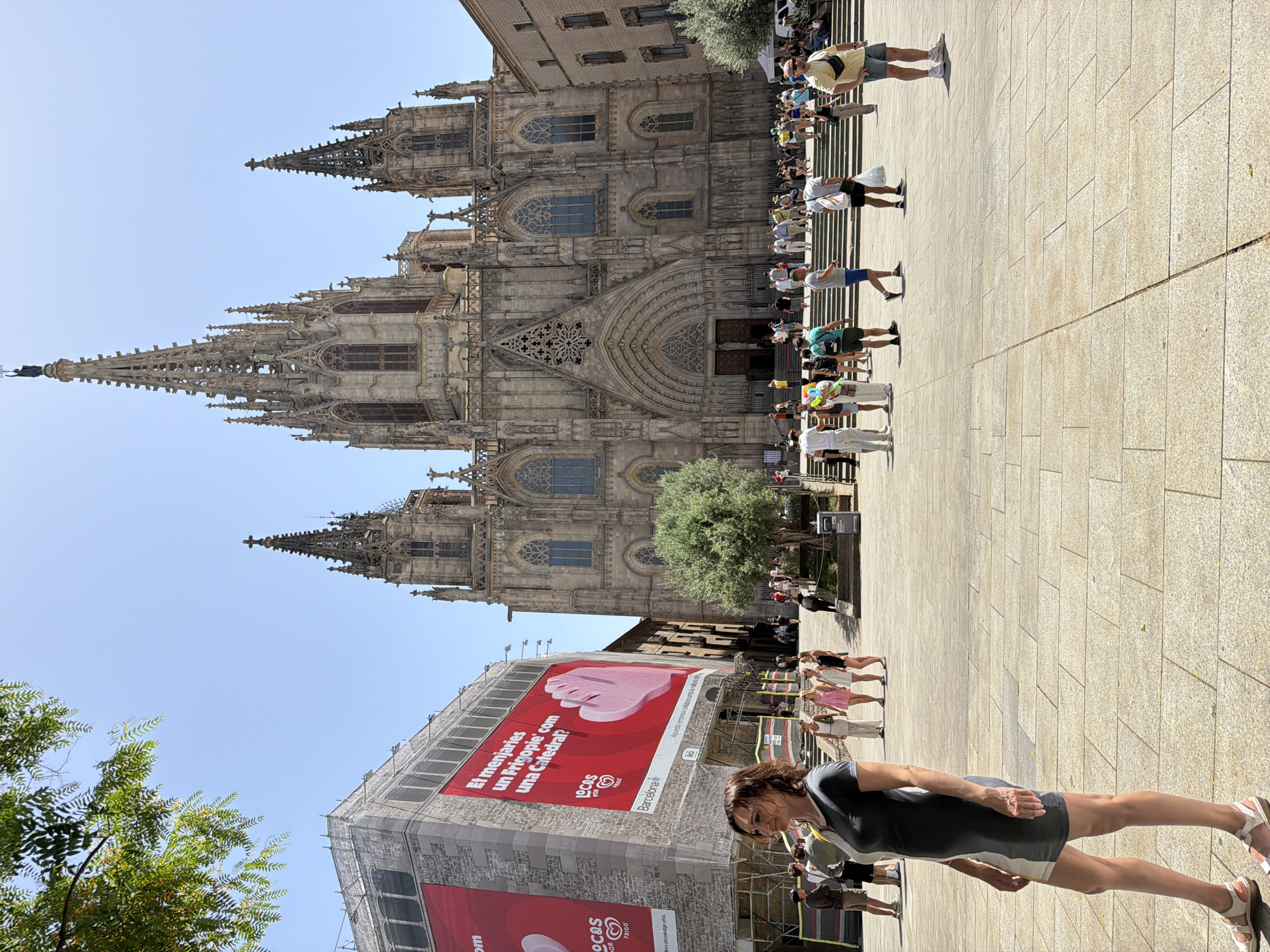 The image depicts the Barcelona Cathedral in Barcelona, Spain. In the foreground, a woman stands to the left, facing right and looking to the left. She is wearing a two-toned short-sleeved dress and sandals. Behind her, several other people are visible, including a group of three women in dresses and a man in shorts and a t-shirt. The background features the imposing Gothic facade of the Barcelona Cathedral with its spires and arched entrances. To the left of the cathedral, a large building is covered in scaffolding and a red banner with text. The ground is paved with large stone tiles. The sky is clear and blue. The scene is illuminated by bright sunlight, casting shadows on the ground. Several tourists are scattered throughout the plaza, some walking and others standing.