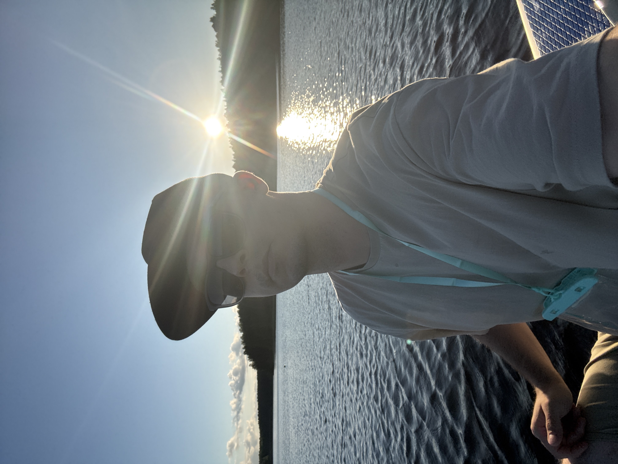 The image captures a selfie taken by a person wearing a dark baseball cap and sunglasses, positioned on a body of water, possibly a lake. The sun is setting behind a tree-lined horizon, casting a bright glare and lens flare across the sky and water. The person is wearing a light-colored t-shirt with a blue lanyard and a clear pouch around their neck. The water is rippled, indicating a slight breeze. The background shows a dense line of trees along the far shore. The scene appears to be outdoors in a natural setting during golden hour. While the specific location of Gora (Khokhlovskoe S/P), Russia, is mentioned, there are no visual cues within the image to confirm this location.