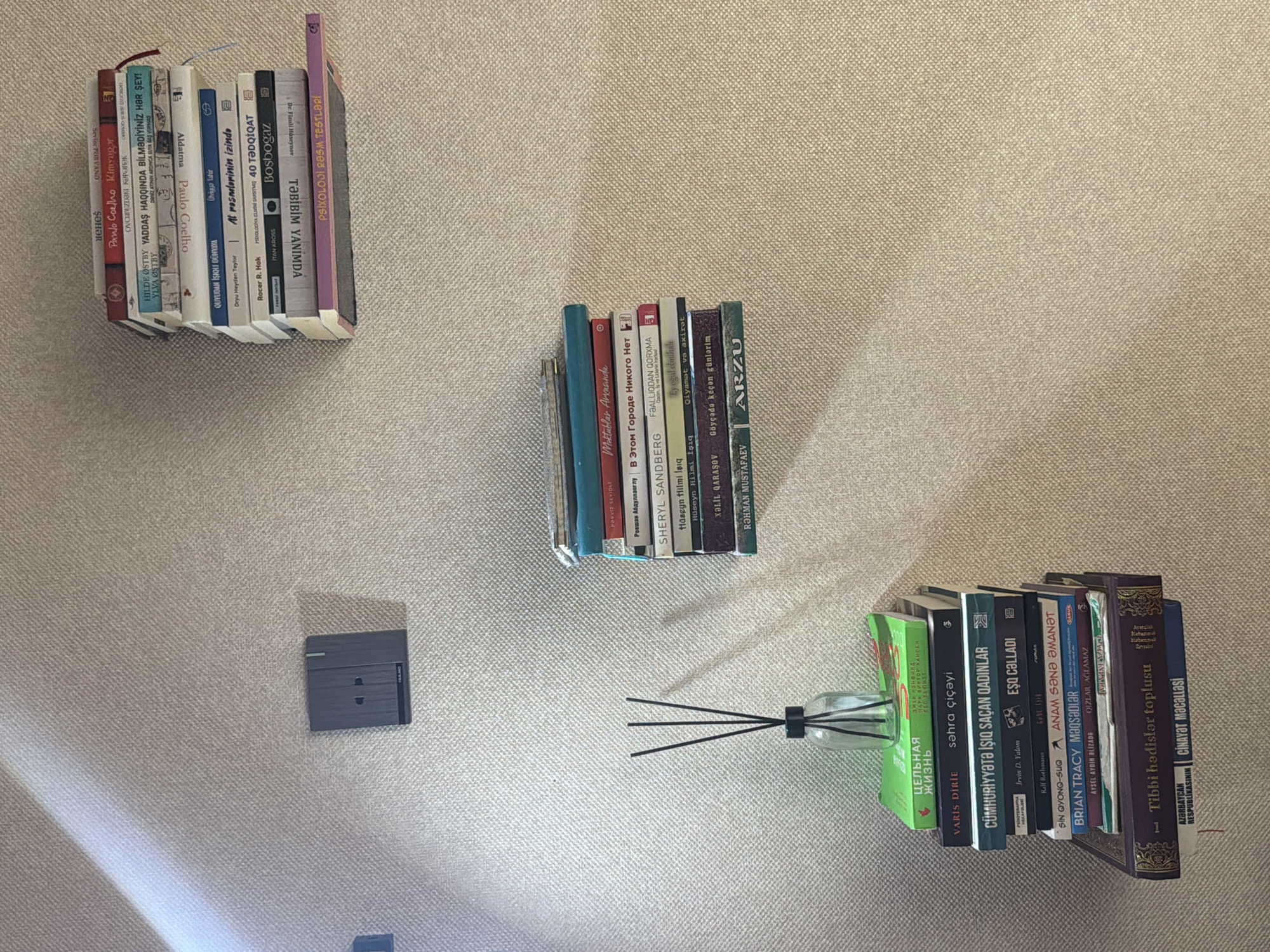 The image shows a wall decorated with two stacks of books and a diffuser with sticks in a bottle. One stack of books is on a shelf on the left side of the wall, next to a wall outlet. The other stack of books is on a shelf on the right side of the wall. The books appear to be a mix of hardcover and paperback, with titles visible in both English and what appears to be Azerbaijani. The wall is textured and beige, with a subtle pattern. A shadow from a light source is visible in the upper left corner of the image. The scene appears to be indoors, possibly in a home or office. The specific location in the city of Sabirabad, Azerbaijan, cannot be determined from the image alone, as there are no identifiable landmarks or contextual clues related to that specific location.