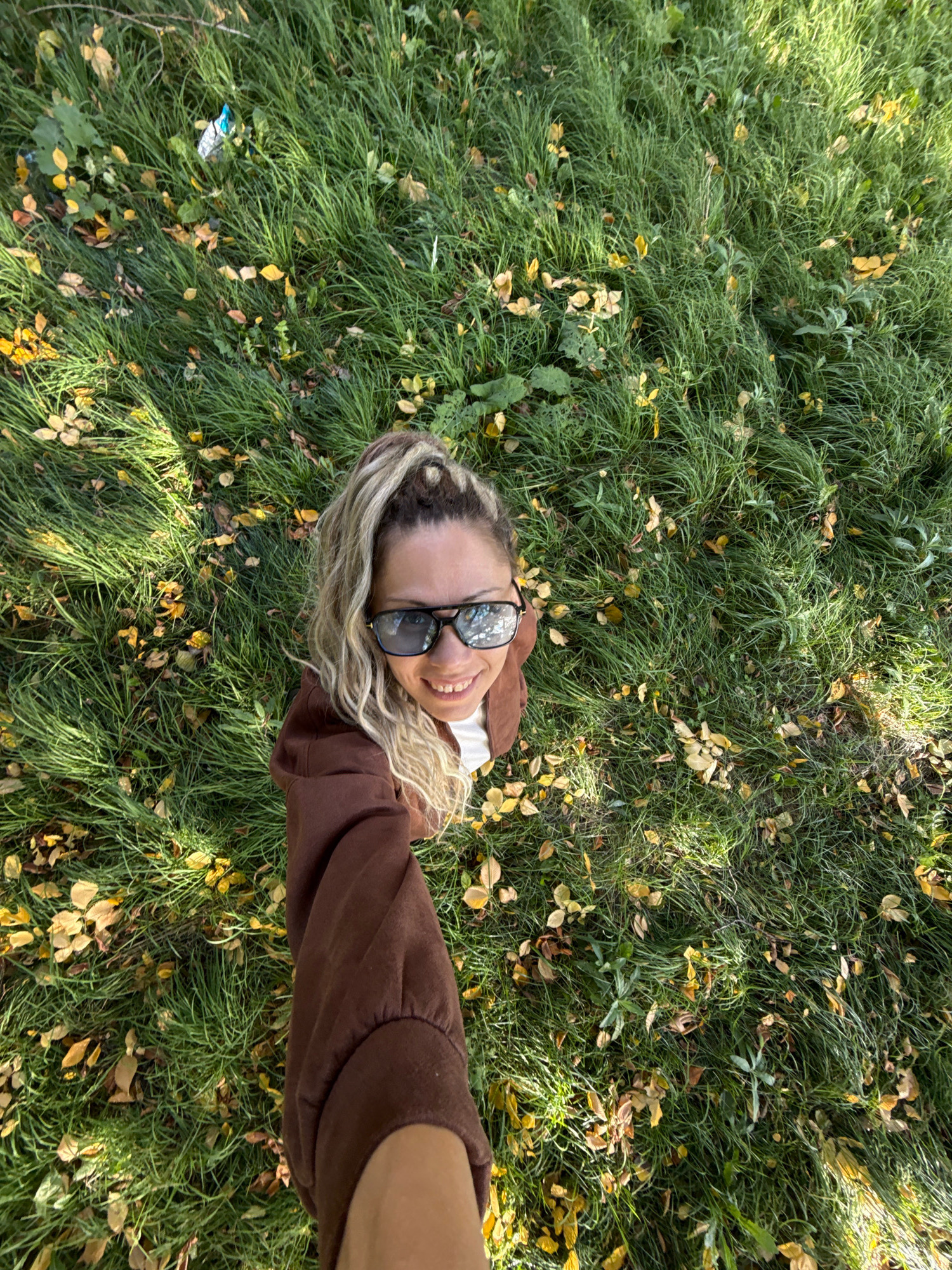 In Rusanovo, Russia, a woman with blonde hair in a ponytail, wearing sunglasses and a brown hoodie, smiles at the camera while standing on grass with fallen leaves. The sunlight casts shadows on the ground. In the upper left corner, a piece of trash is visible amidst the grass and leaves.