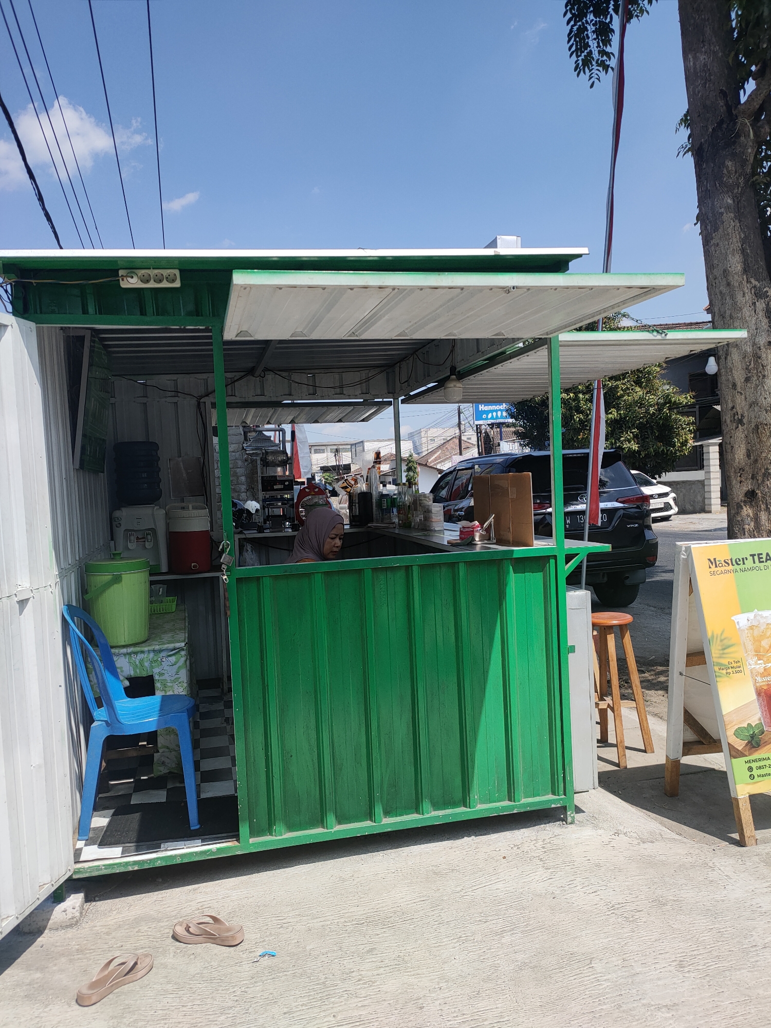 A small, green food stall is set up on a concrete sidewalk in front of a tree. A woman wearing a hijab is visible inside the stall, serving customers. A blue chair is placed outside the stall, and a pair of flip-flops lies on the ground. A sign in front of the stall advertises "Master Tea." In the background, a black SUV and a white car are parked on the street. The sky is clear and blue, with a few wispy clouds. The scene appears to be in the city of Kanigoro, Indonesia.