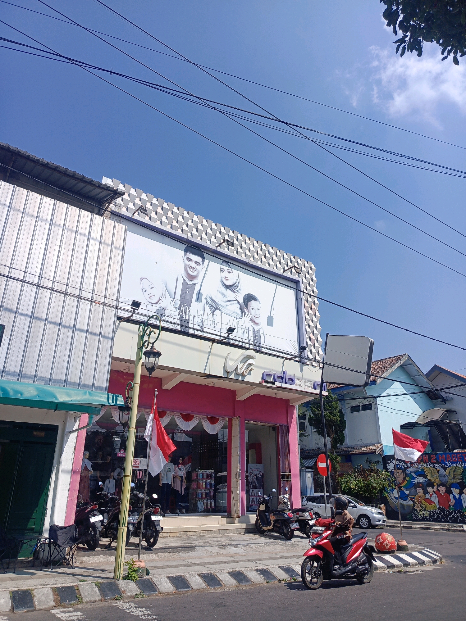 The image captures a street view in Sukowinangun, Indonesia, on a clear day. A storefront with a large billboard featuring a family is prominently displayed. The billboard is framed with a geometric pattern. Below the billboard, the store has a pink facade and large glass windows revealing mannequins dressed in clothing. Two motorcycles are parked outside the store. A person is riding a red motorcycle in the foreground, moving from right to left across the frame. The street is paved with asphalt and has a pedestrian crossing. Overhead, a tangle of electrical wires crisscrosses the sky. To the right of the storefront, another building with a light blue facade is visible, adorned with a mural depicting people and a flag. A red and white Indonesian flag flies on a pole near the mural. Trees and foliage are present in the background.