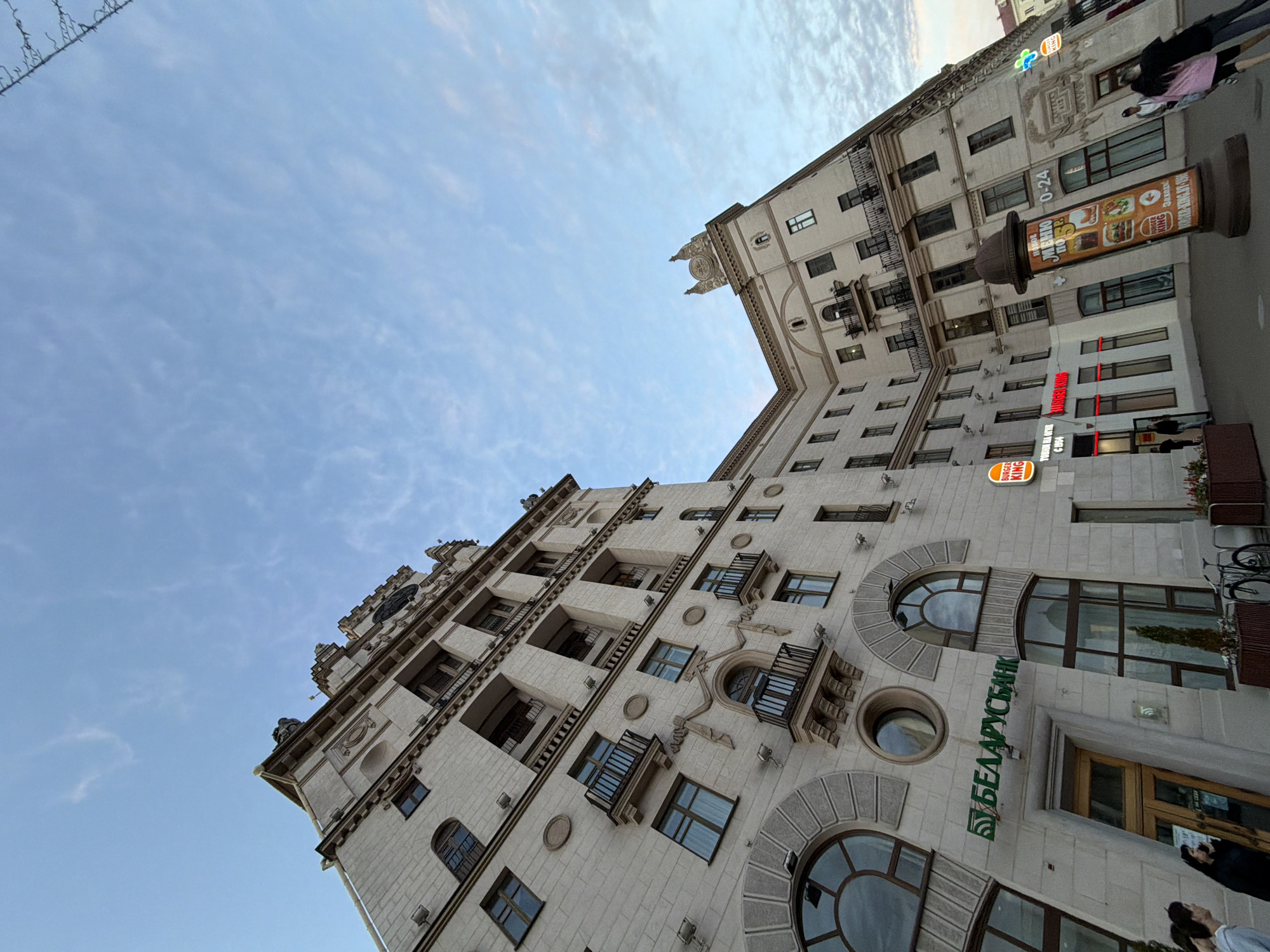 An upward-looking, tilted perspective shot captures a grand, ornate building in an urban setting, identified as Minsk, Belarus. The multi-story structure is light-colored, featuring numerous windows, some with small balconies and decorative arched surrounds. Its architectural style suggests a classical or Soviet-era influence, with intricate detailing and cornices visible on its facade. The sky above is a clear light blue with scattered wispy white clouds, indicating daytime, possibly late afternoon.
On the ground floor, commercial establishments are prominently featured. A large green sign reads "БЕЛАРУСБАНК" (Belarusbank). Higher up, a red "BURGER KING" sign is visible, accompanied by a tall cylindrical advertising column displaying Burger King promotions. Text on the column includes "МЕНЮ 0-24" (Menu 0-24), "ТОВИМ НА ОГНЕ" (Cooking on fire), "C1954", and "BURGER-KING.BY-1964". Another smaller Burger King logo appears towards the top right. Several people are visible on the sidewalk below, either walking or standing, and what appears to be a bicycle rack or parked bicycles is also present. The dynamic angle emphasizes the building's height and decorative elements against the expansive sky.
