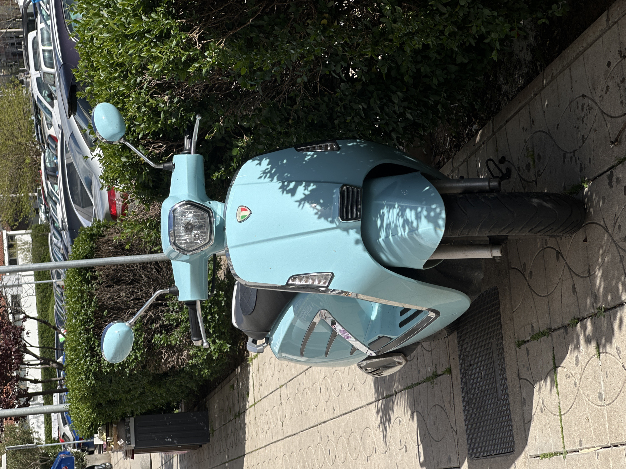 A light blue moped is parked on a patterned sidewalk in Šušanj, Montenegro. Green shrubs surround the moped. Cars are parked in the background.
