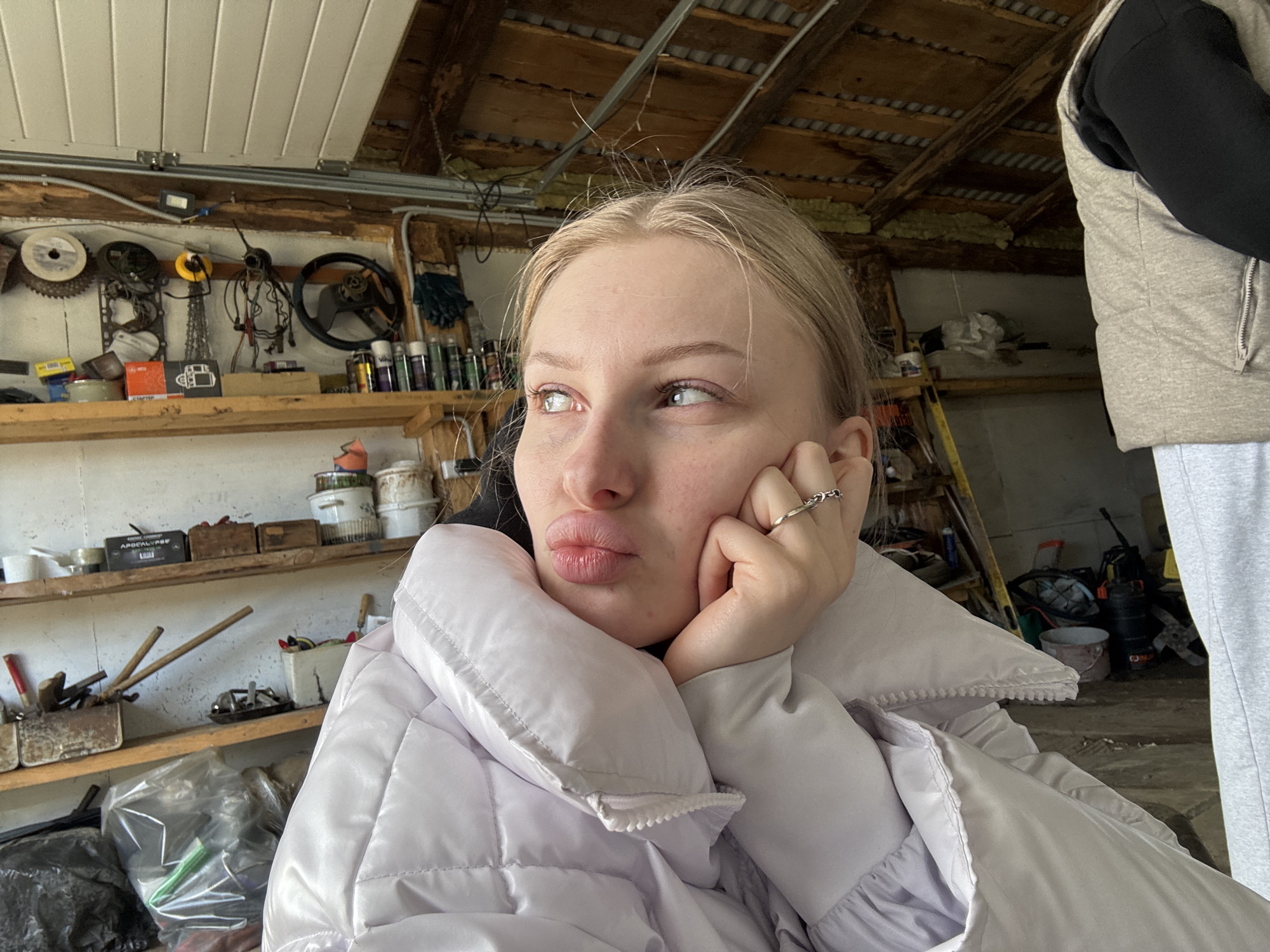 A woman with fair skin and blonde hair is pictured indoors in what appears to be a garage or workshop. She is wearing a light purple puffer jacket and a ring on her finger. She is resting her cheek on her hand, looking off to the side. The background shows shelves with tools and equipment, a steering wheel hanging on the wall, and a wooden ceiling. Part of another person is visible on the right side of the frame, wearing a grey vest and sweatpants. The location is potentially Бородулино, Russia.