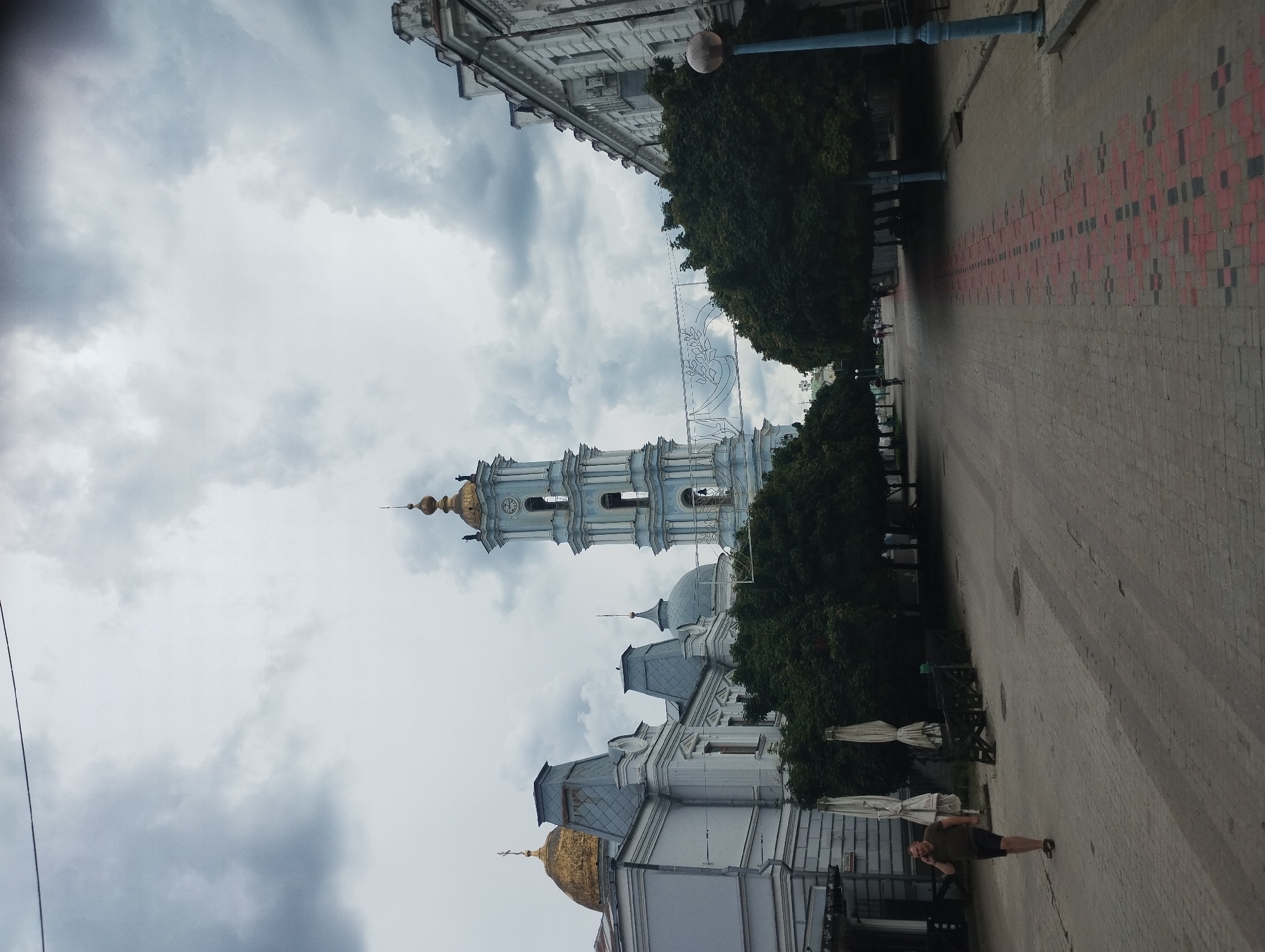 The image depicts a view looking down a paved pedestrian street in Sumy, Ukraine, towards a prominent bell tower. The sky is overcast with dramatic, dark clouds. On the left side of the street, a light-colored building with a golden dome and ornate architectural details is visible, adjacent to a blue and white bell tower that dominates the central background. The bell tower features multiple tiers and arches, with a clock face on one level. A decorative white banner, possibly for an event, is strung across the street between the bell tower and a building on the right.
On the right side of the street, a white building with many windows and classical architectural elements lines the pedestrian walkway. The walkway itself is paved with cobblestones, with a decorative pattern of red and dark square tiles forming a central border. Lush green trees line both sides of the street, casting shadows on the pavement.
In the foreground on the left, a man wearing a dark green t-shirt and dark shorts is walking away from the camera while talking on his cell phone. He is wearing sandals. Behind him, several closed white umbrellas are visible, suggesting a cafe or outdoor seating area. Further down the street, other pedestrians and a cyclist can be seen in the distance. A blue lamppost stands on the right side of the walkway. The overall impression is of a historic urban environment under a moody sky.