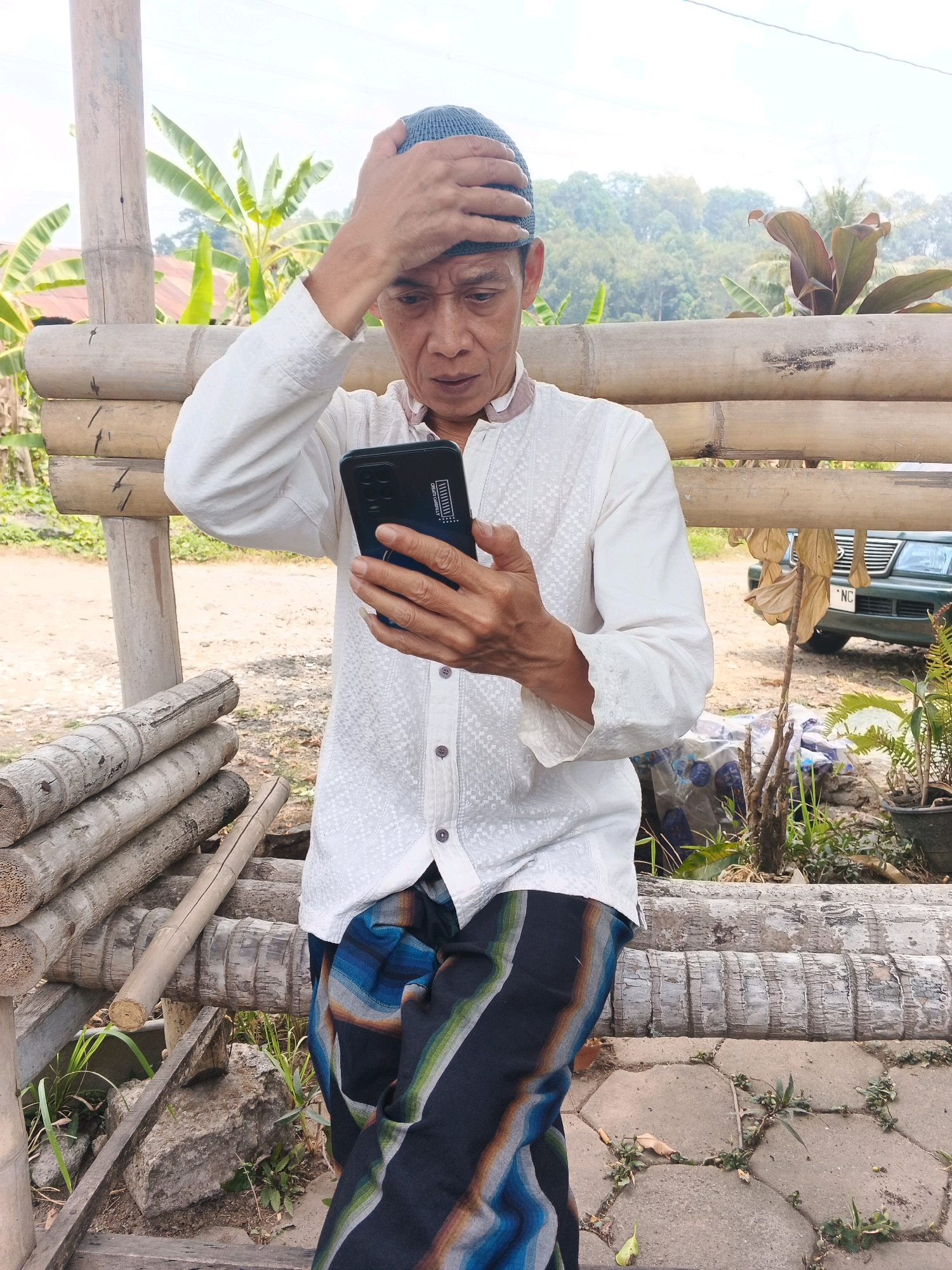 A middle-aged man wearing a white embroidered long-sleeved shirt and a blue skullcap, with a patterned sarong tied around his waist, stands outdoors near a structure made of bamboo logs. He is holding a black smartphone in his right hand and touching his forehead with his left hand, appearing concerned or thoughtful. The background features banana trees, foliage, and a green car with a license plate that appears to read "NC". The scene is set on a concrete or paved surface with some weeds growing between the paving stones, suggesting an outdoor area, possibly a yard or a rural setting in Ungaran, Indonesia.