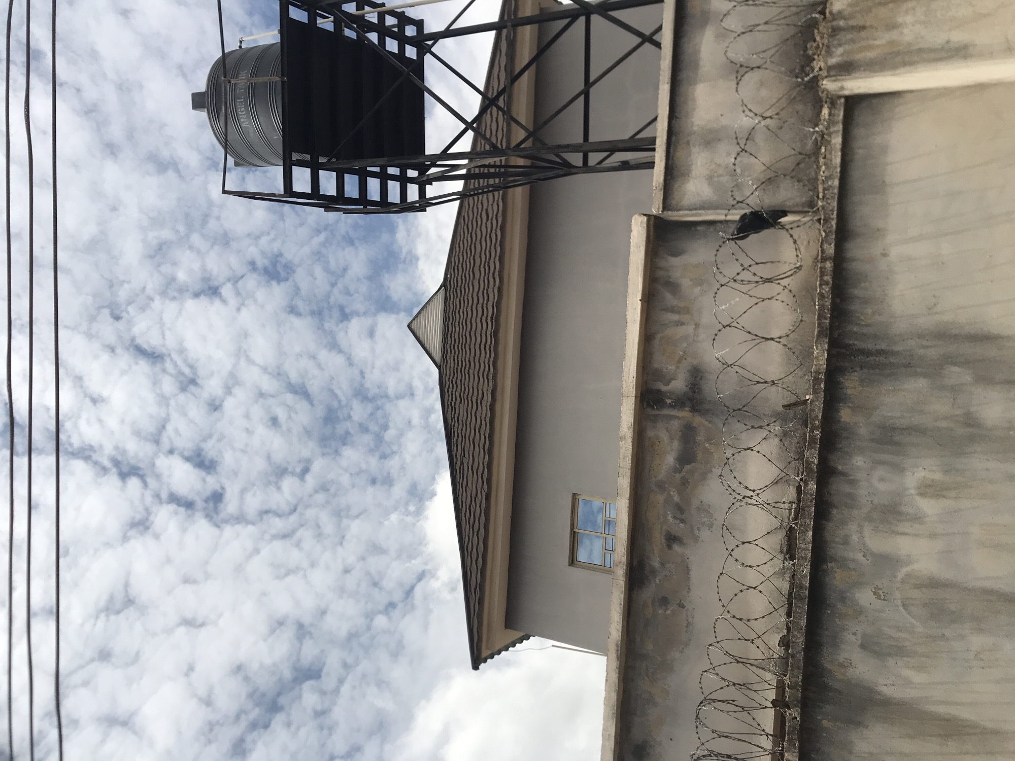 This image captures an outdoor scene in Rafin Par, Nigeria, viewed from a low angle. The upper portion of the frame is dominated by a sky filled with scattered white cumulus or stratocumulus clouds against a light blue background. Three dark, horizontal utility lines traverse the top of the image. A two-story building with smooth, light grey walls and a dark brown, corrugated hip roof is centrally positioned, with a small, rectangular window on its upper facade reflecting the cloudy sky. To the right of the building, a large, dark grey cylindrical water tank, bearing the label "ANGEL TANK," is elevated on a tall, black metal stand with an open lattice structure. The lower section of the image shows a weathered concrete wall, stained and discolored, topped with a continuous line of coiled razor wire. In the lower right corner, a dark, indistinct object, possibly a piece of fabric or a bag, is draped over the wall, partially entangled with the razor wire.