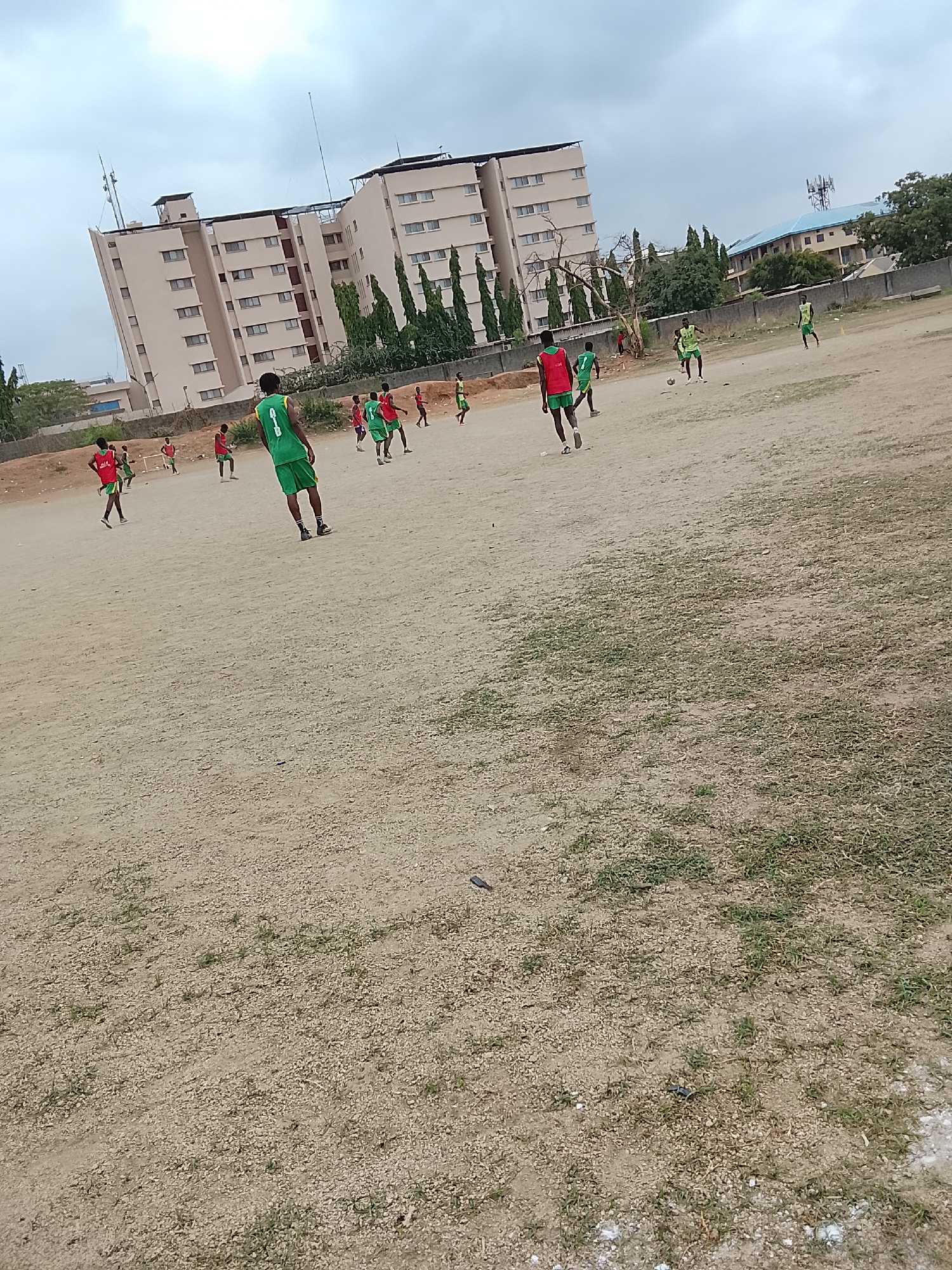 A soccer game is in progress on a dirt field in Abuja, Nigeria. Approximately twelve players wearing green and red jerseys are scattered across the field. A multi-story beige building with numerous windows is visible in the background, as well as a blue-roofed building in the distance. The sky is overcast. The field is mostly dirt with patches of sparse grass.